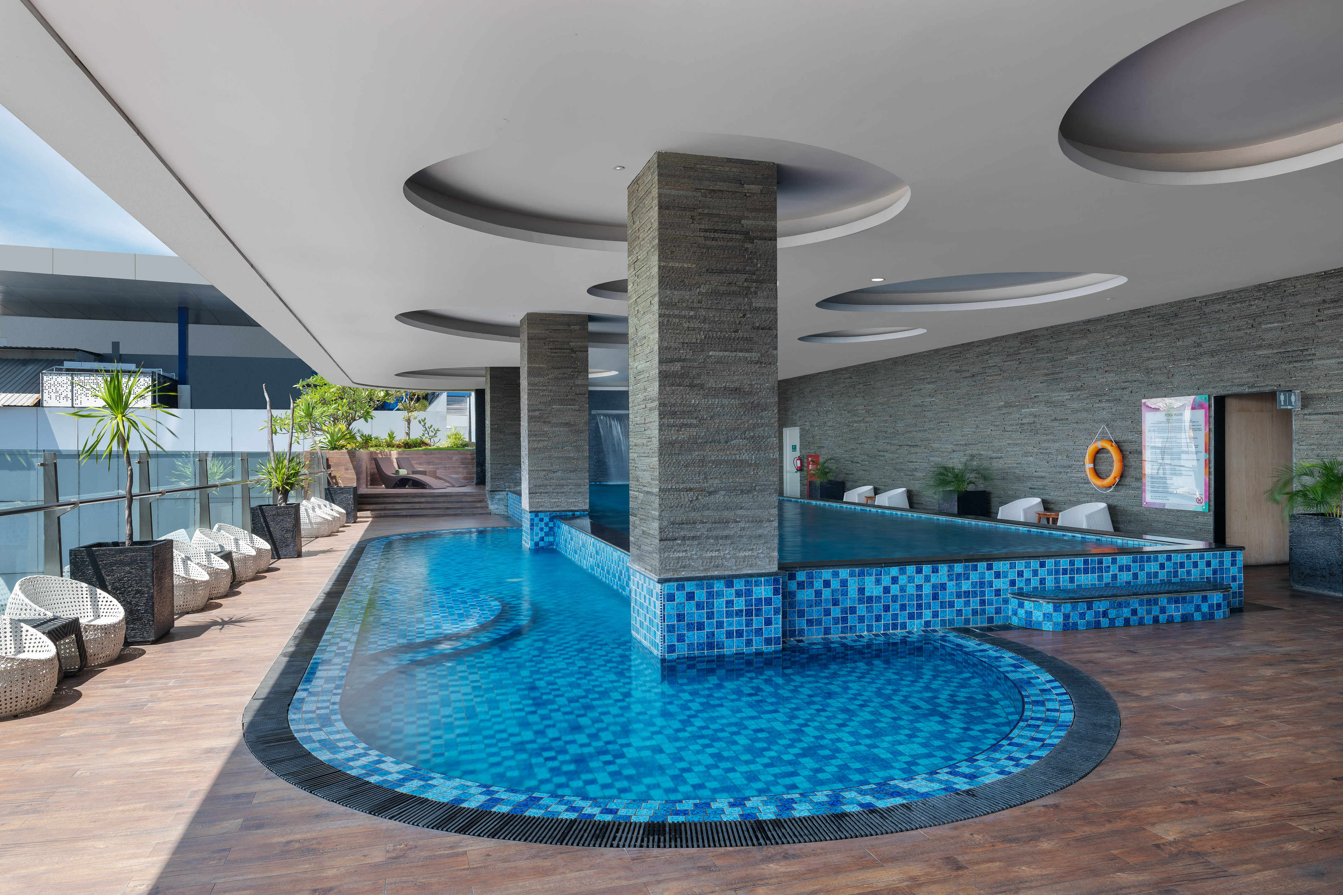 Modern resort swimming pool with blue tiles, wooden deck, and stone pillars.