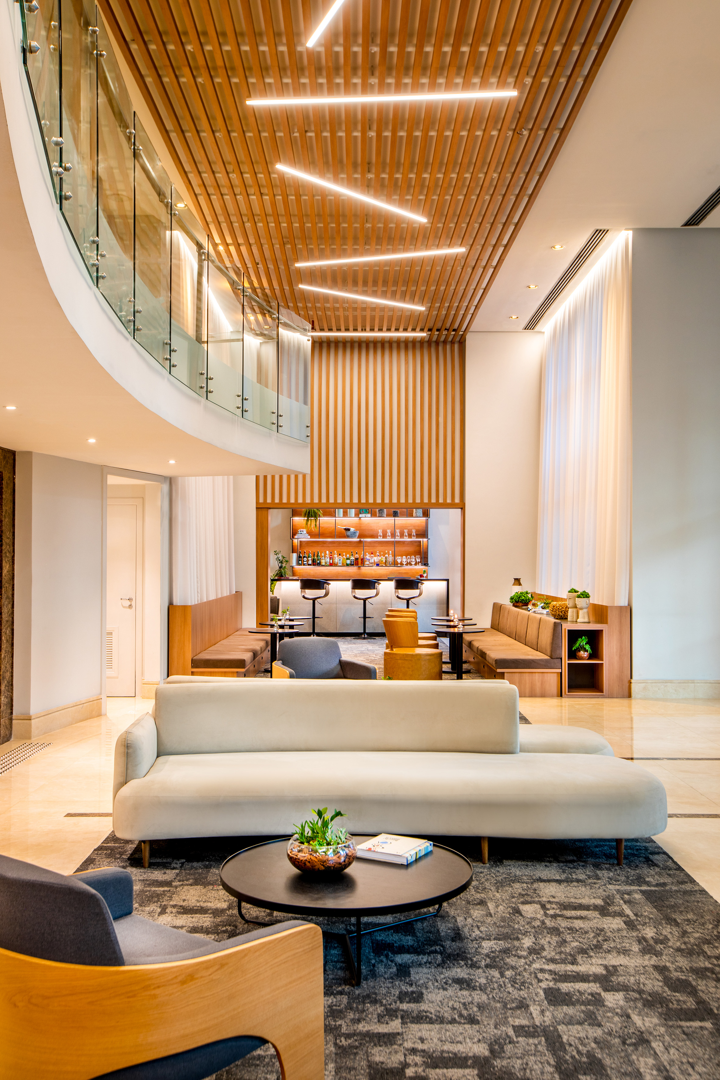 Modern hotel lobby with wood ceiling, glass railing, bar, and plush seating.