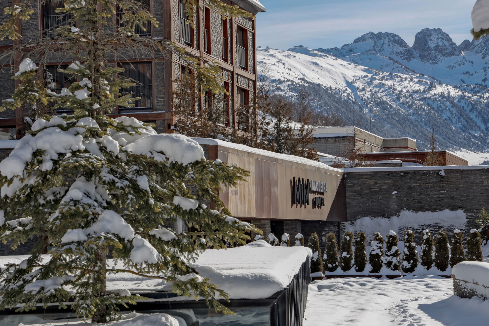 MIM Baqueira Hotel in winter, snow-covered pine tree, and mountains.