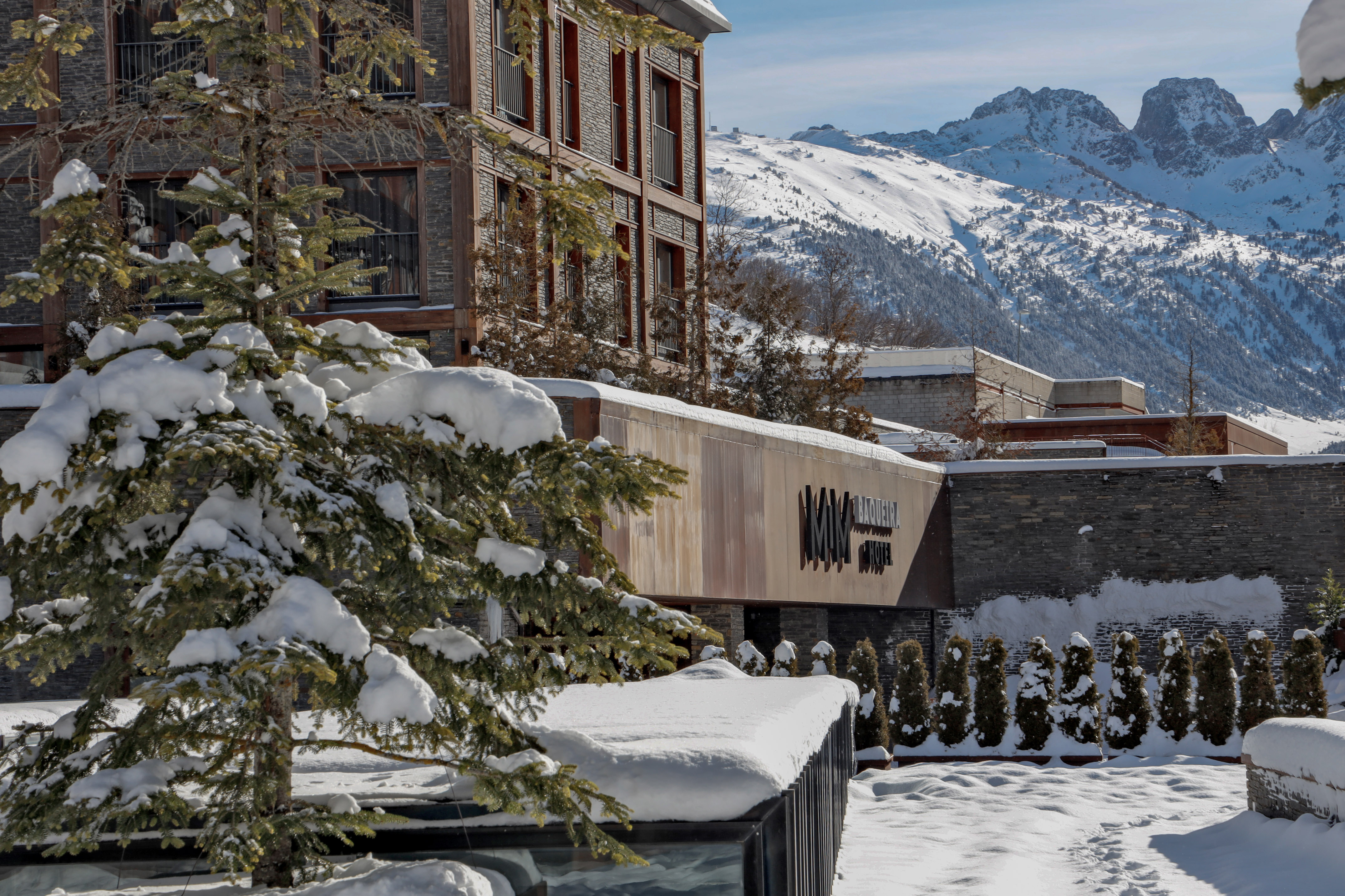 MIM Baqueira Hotel in winter, snow-covered pine tree, and mountains.