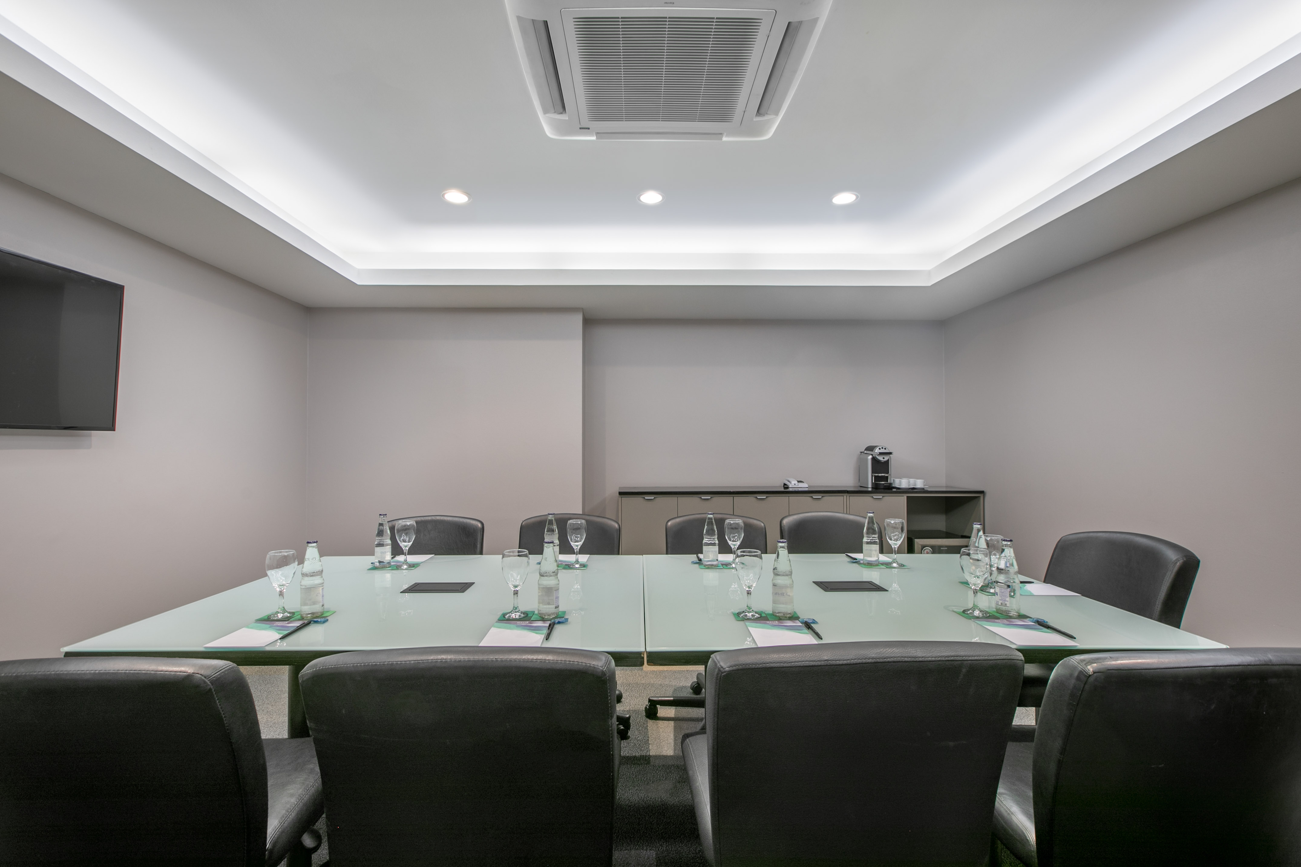 Modern meeting room with glass table, black chairs, TV, and coffee station.