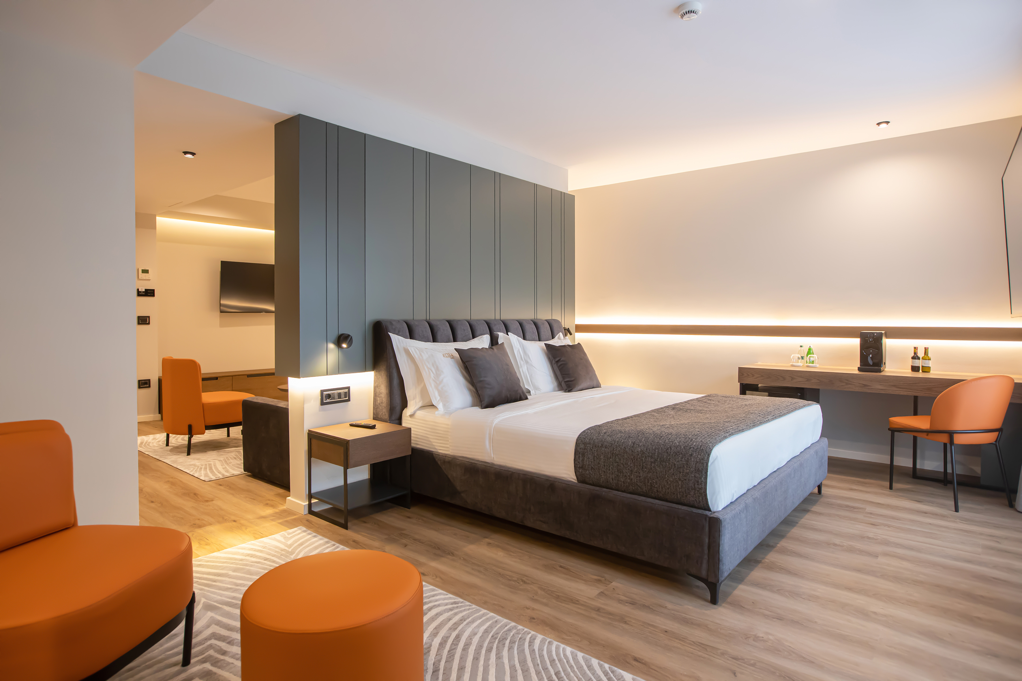Modern hotel bedroom suite with grey upholstered bed and orange seating.