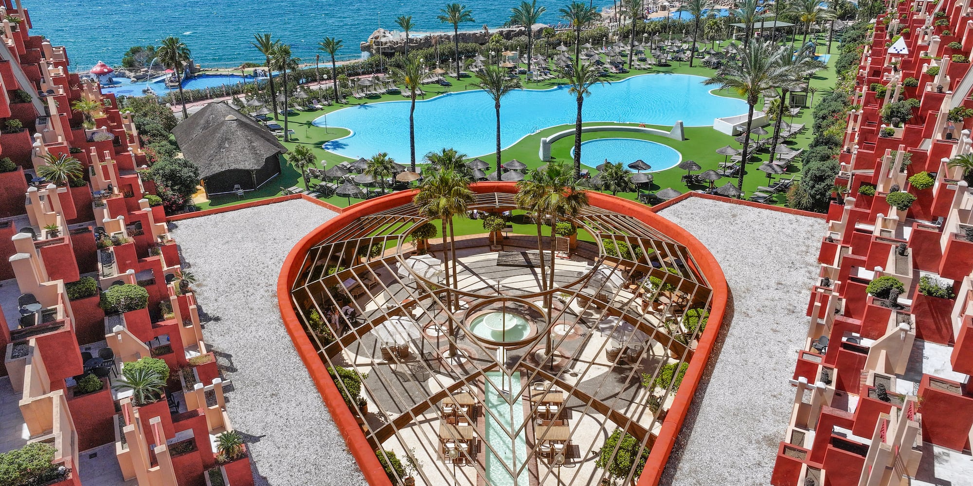 Overhead view of resort, blue pools, palm trees, ocean, and red tiered buildings.