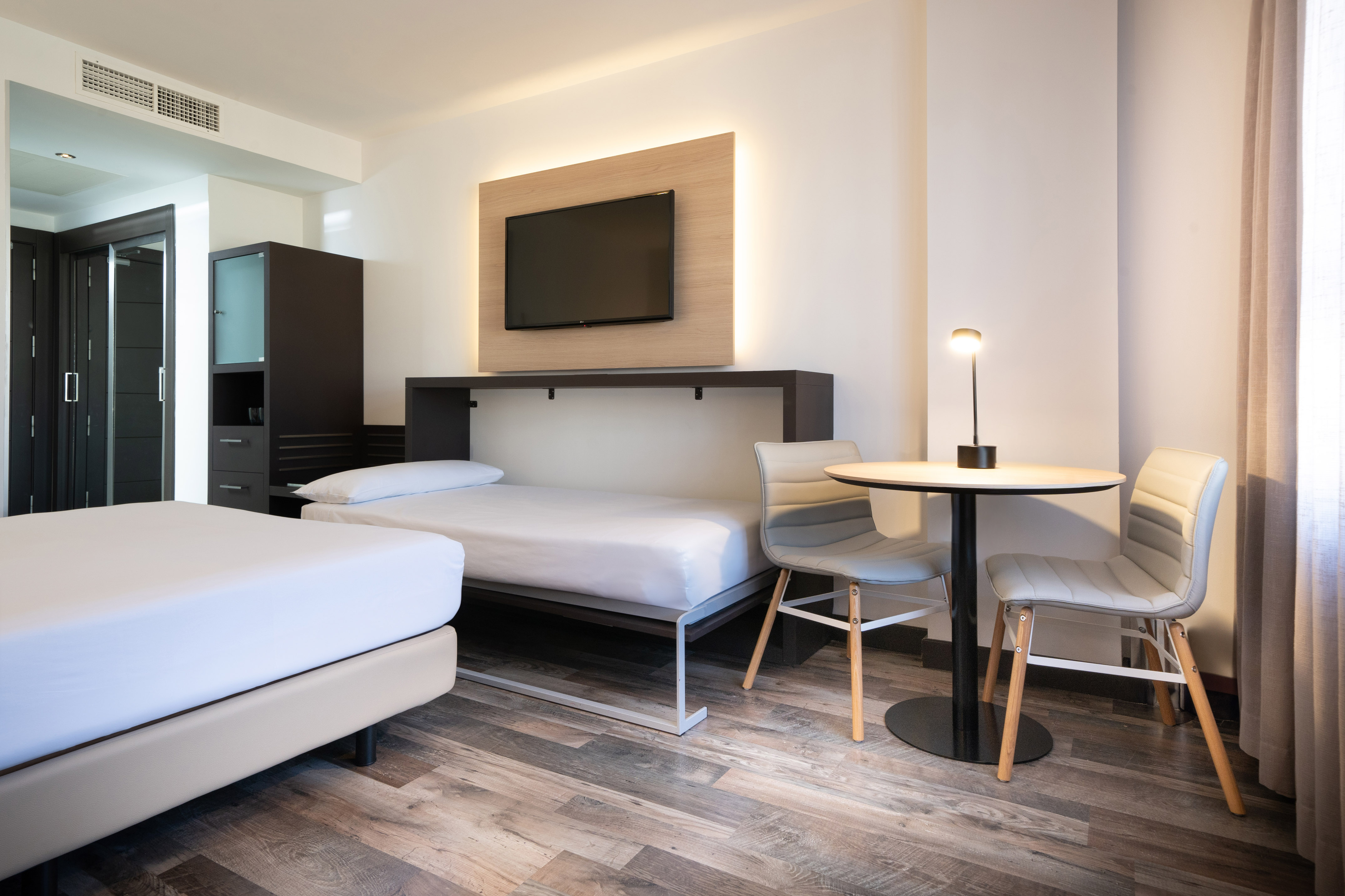 Modern hotel room with twin beds, wall-mounted TV, and small table.