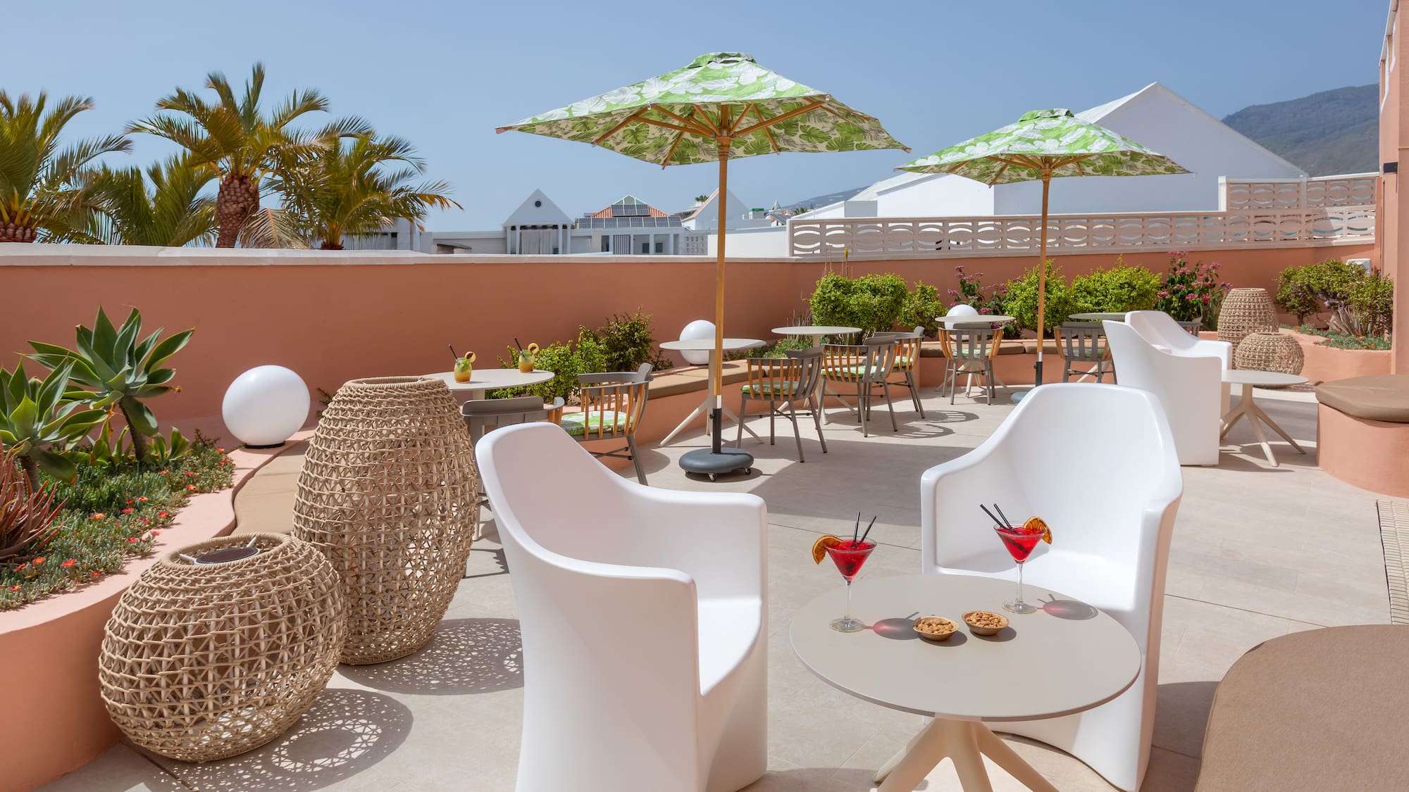 Sunny hotel terrace with modern seating, green umbrellas, cocktails, palm trees.