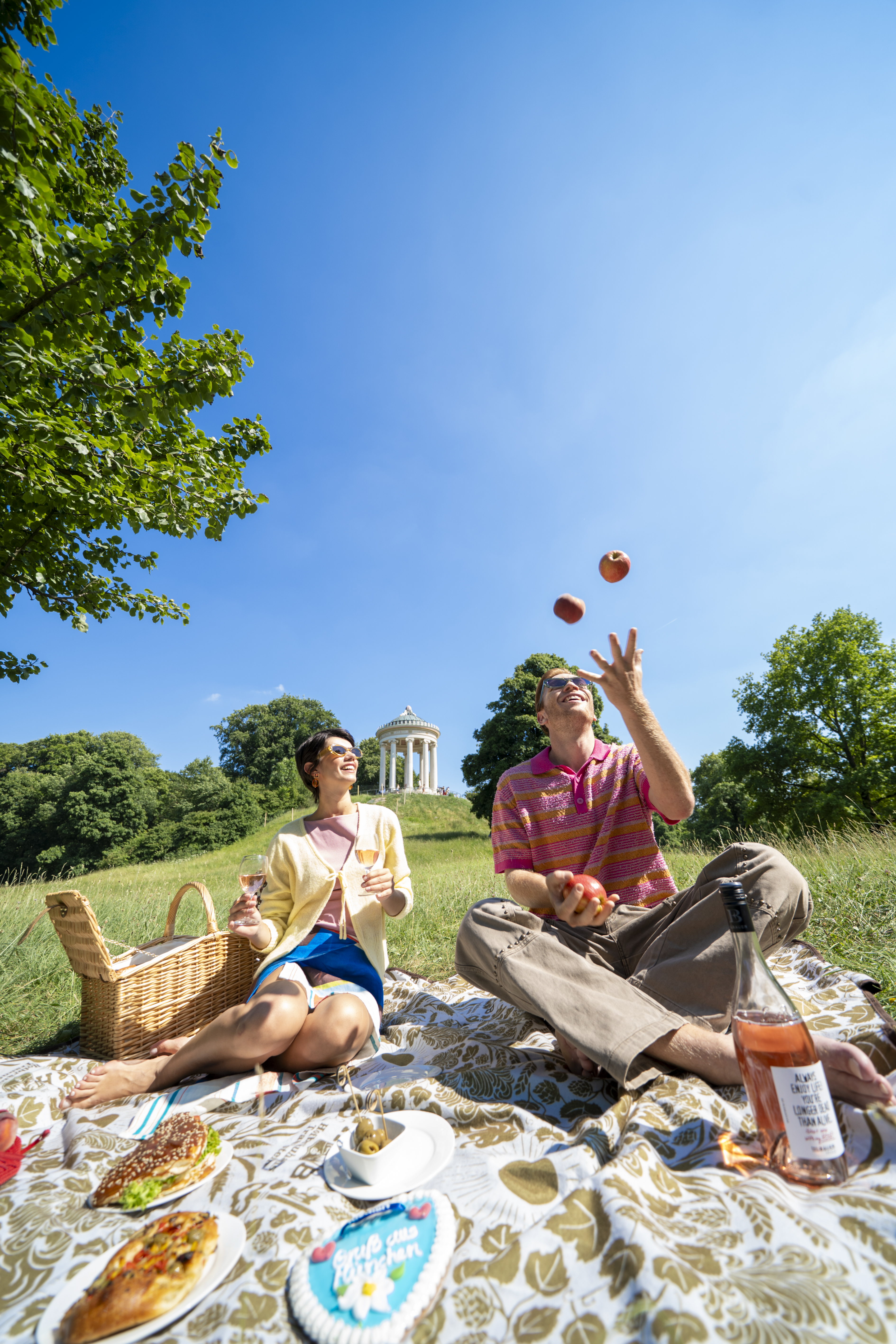Couple picnicking on sunny day, man juggling apples, woman with wine, temple background.