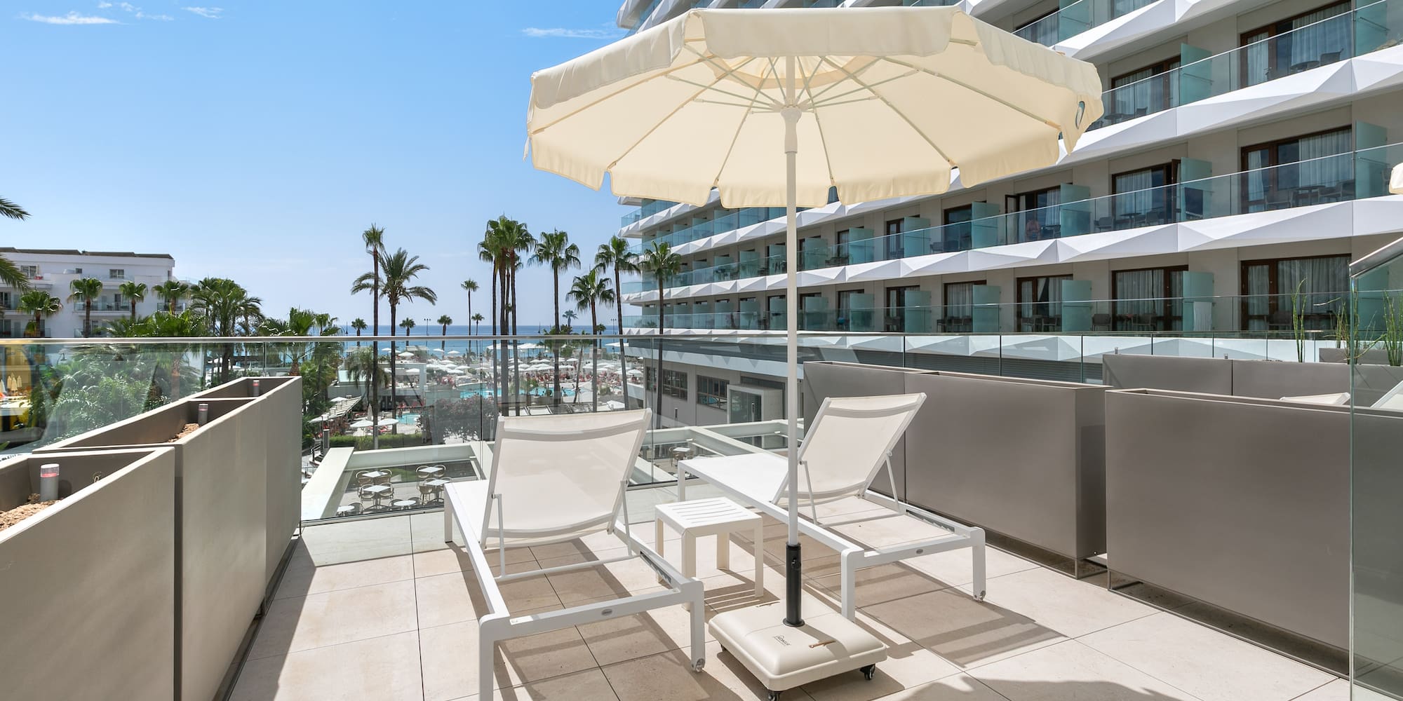 a deck with chairs and umbrella on a balcony with palm trees and a blue sky
