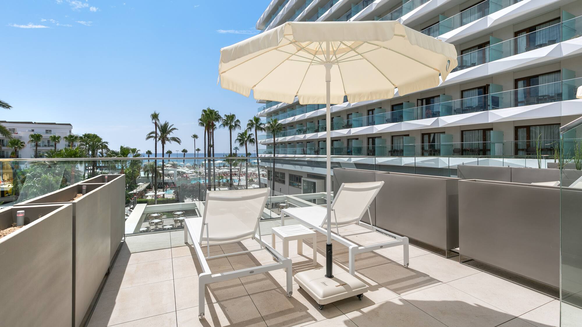 a deck with chairs and umbrella on a balcony with palm trees and a blue sky