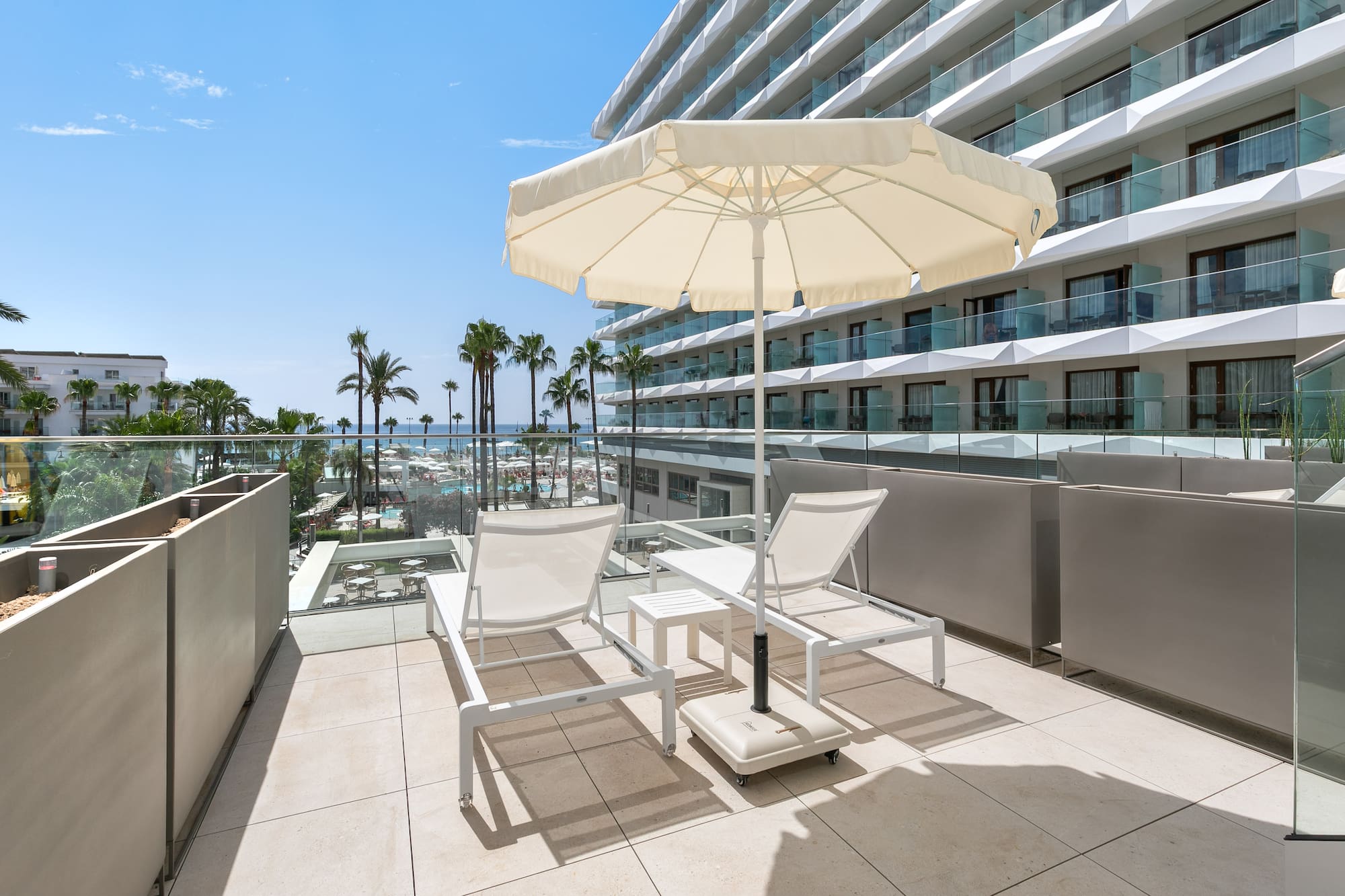 a deck with chairs and umbrella on a balcony with palm trees and a blue sky