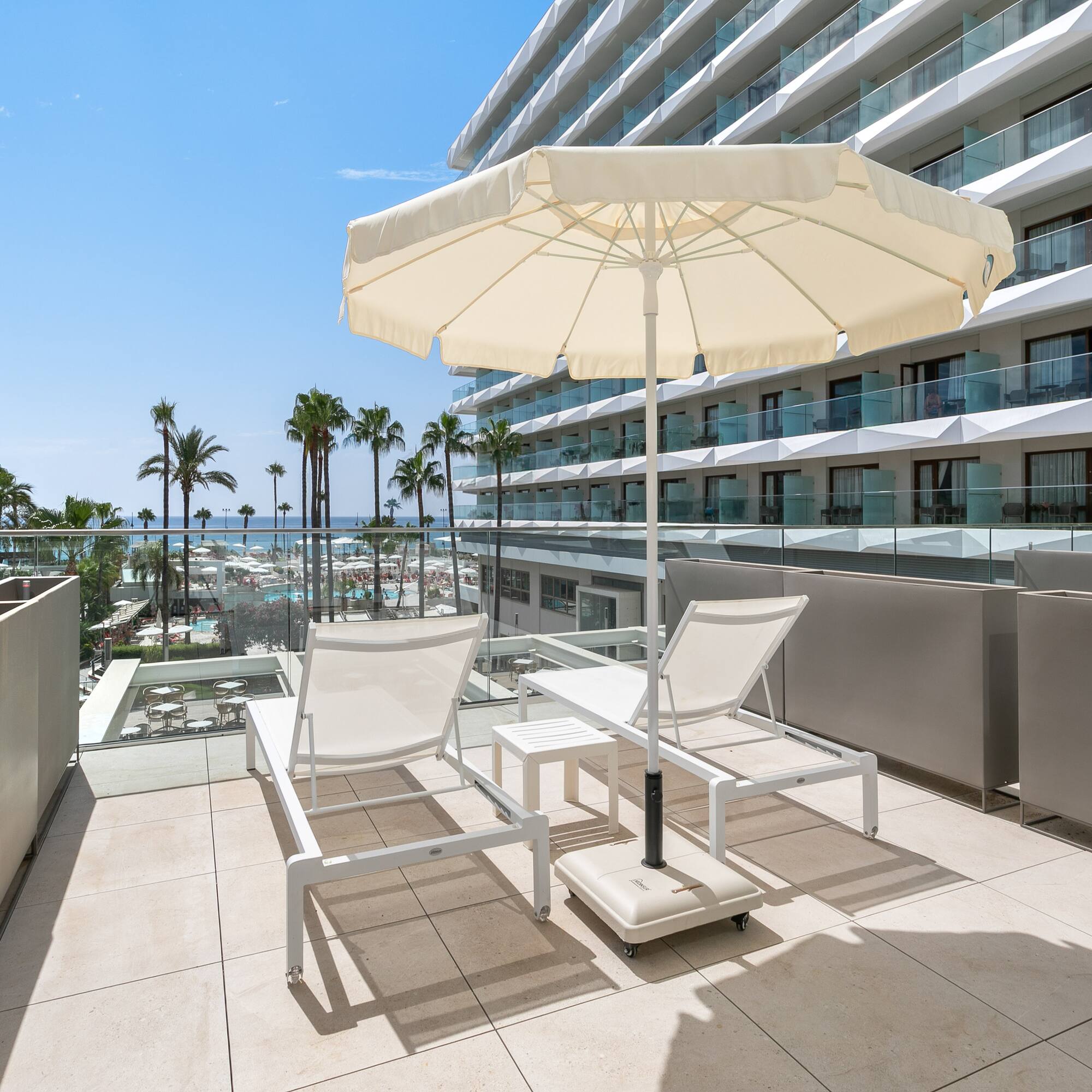 a deck with chairs and umbrella on a balcony with palm trees and a blue sky