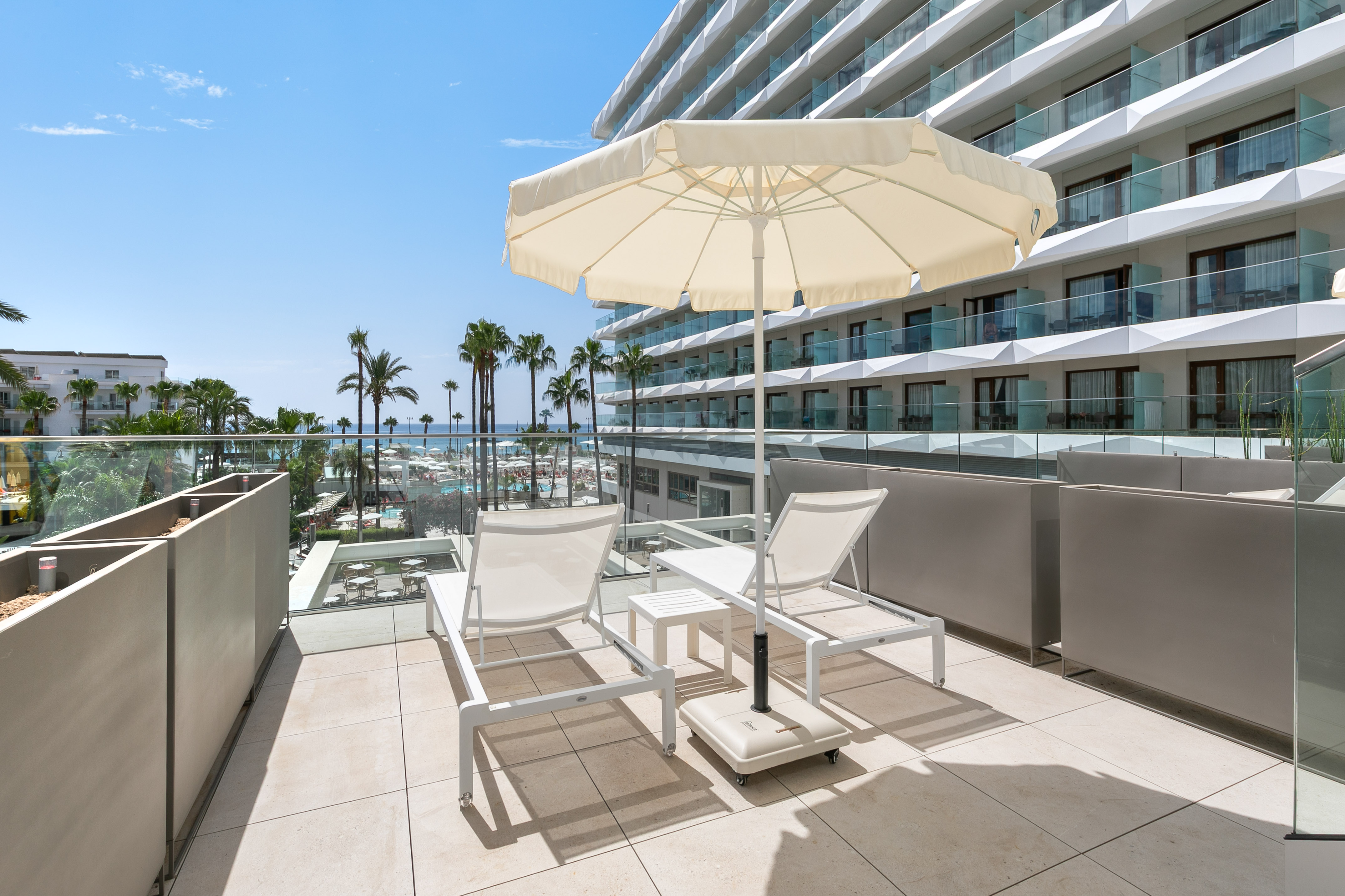 Sunny hotel balcony with lounge chairs, umbrella, palm trees, and sea view.