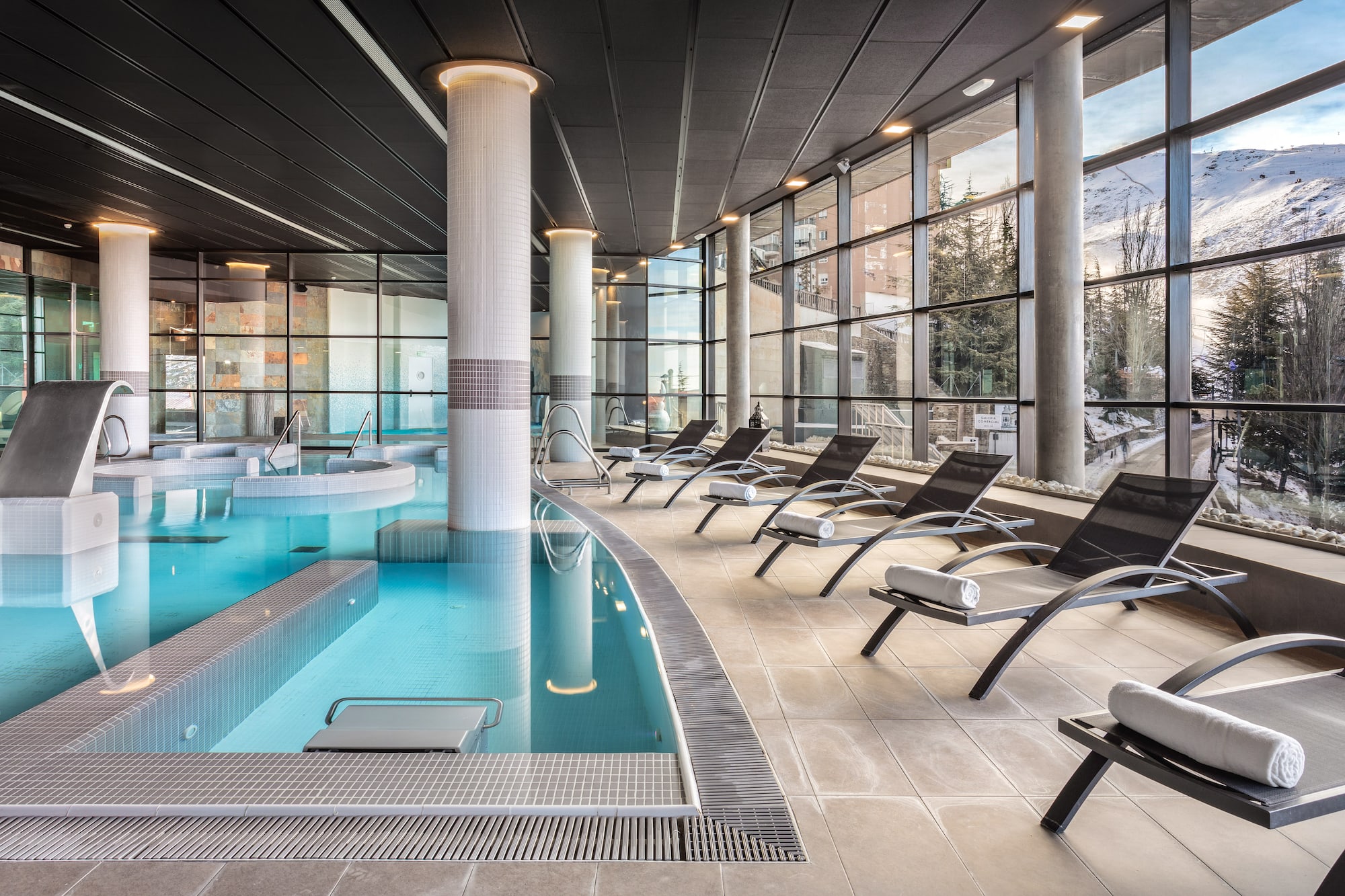 Modern indoor spa pool with mountain views and lounge chairs.