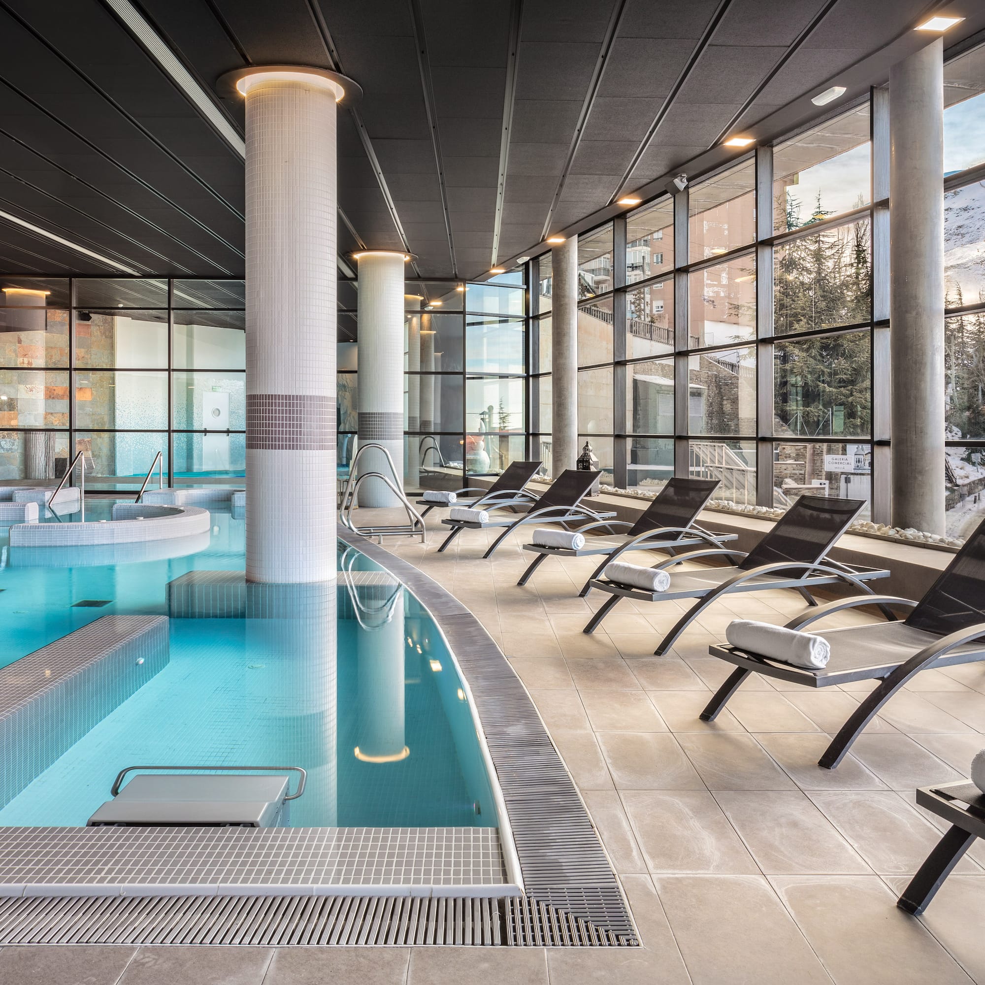 Modern indoor spa pool with lounge chairs and snowy mountain view.