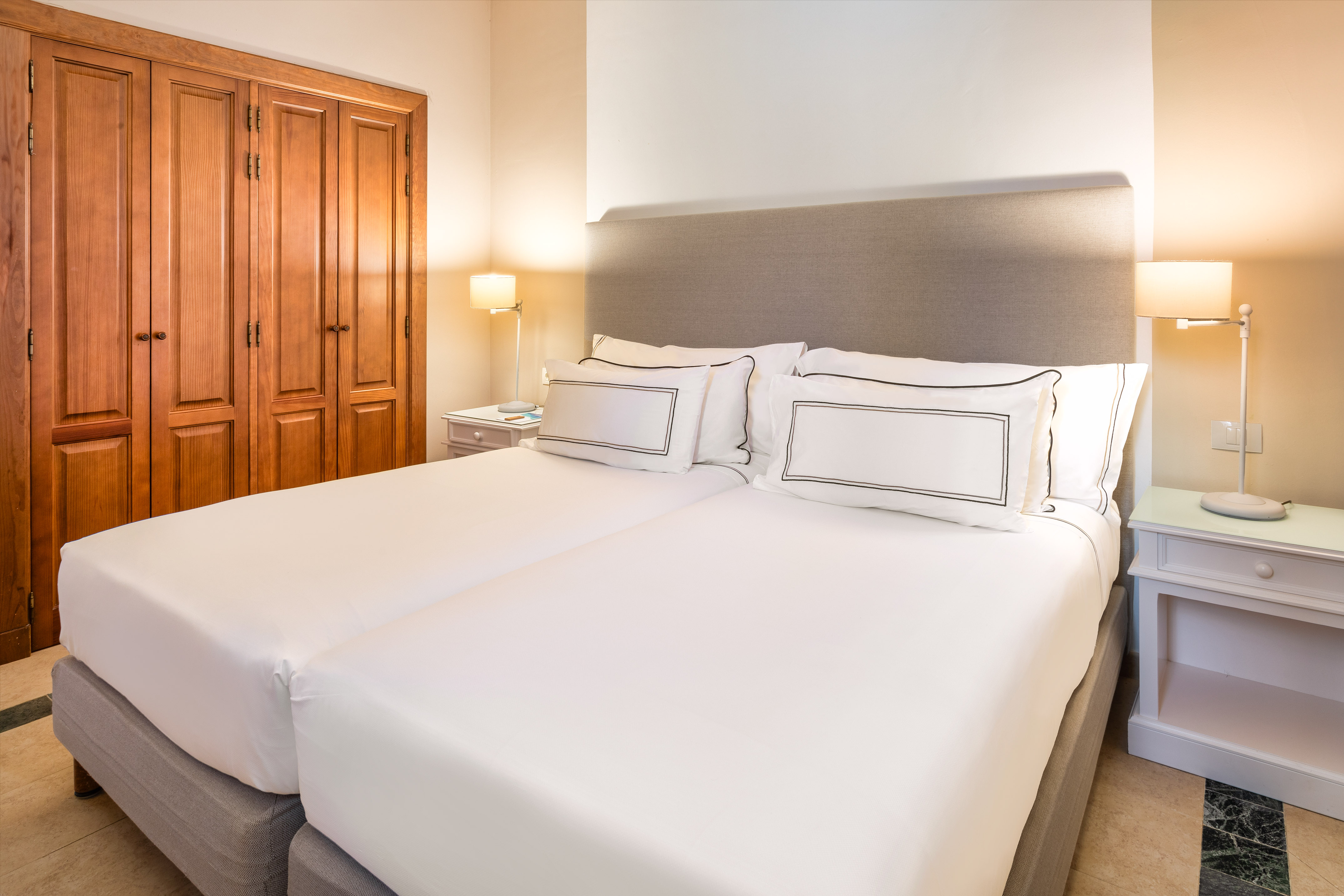 Bright hotel room with twin beds, white bedding, nightstands, and wood wardrobe.
