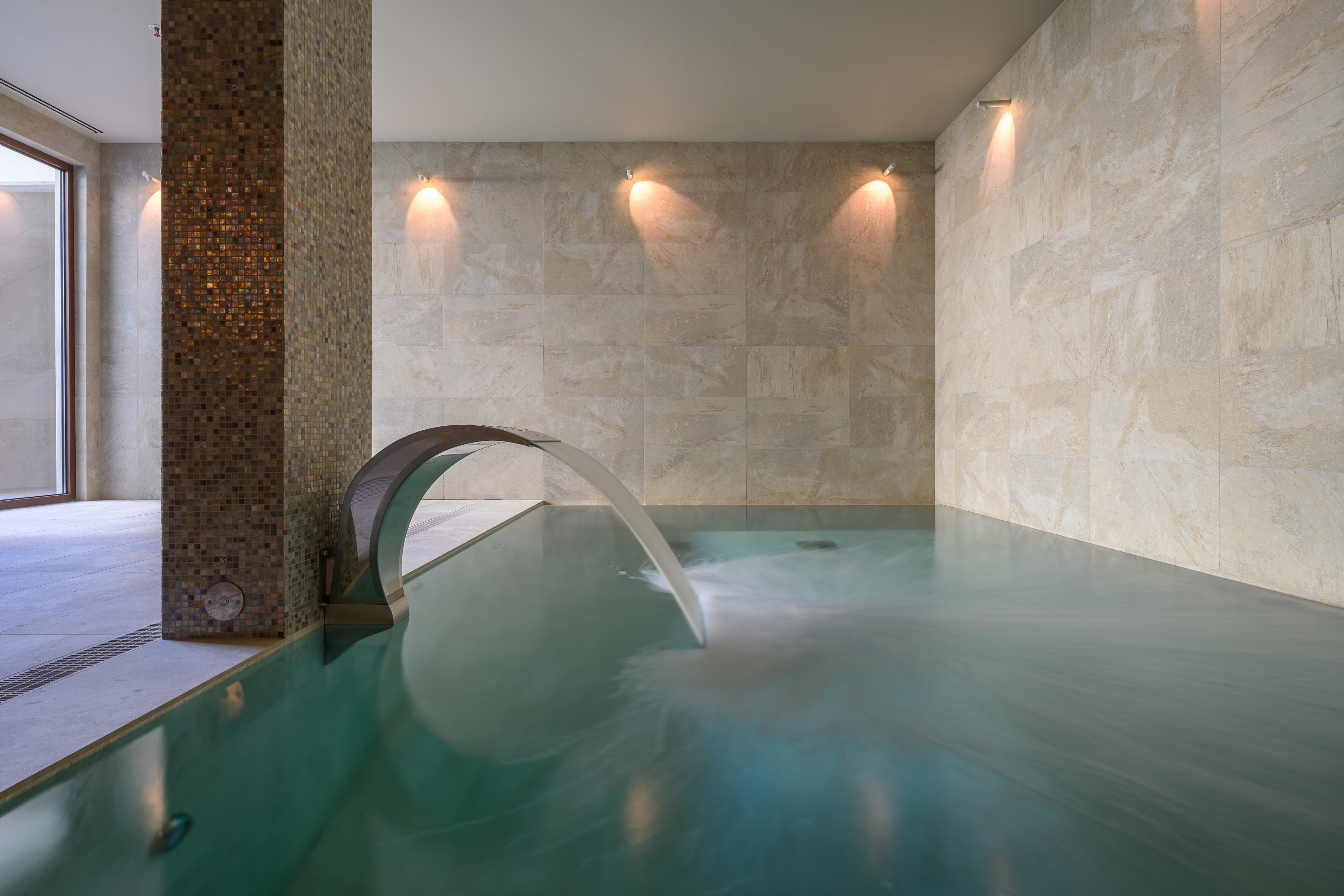 Luxury indoor pool with water feature, tiled walls, mosaic column.