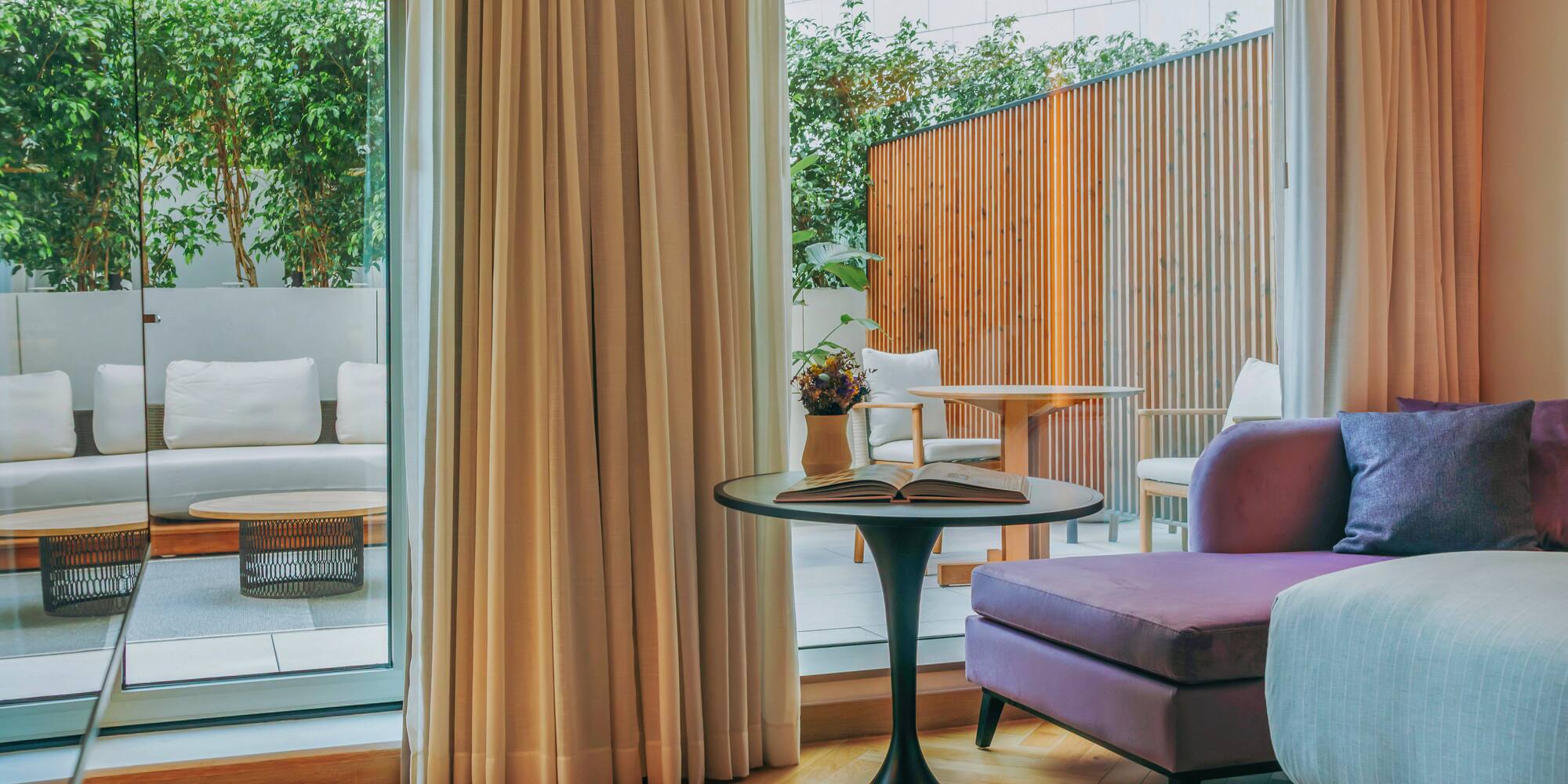 Modern hotel suite with purple sofa, outdoor patio, and lush green plants.