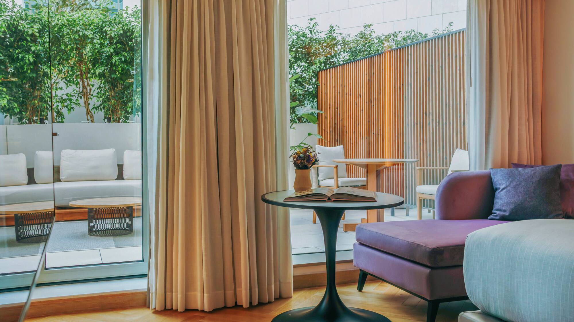 Modern hotel suite with purple sofa, outdoor patio, and lush green plants.