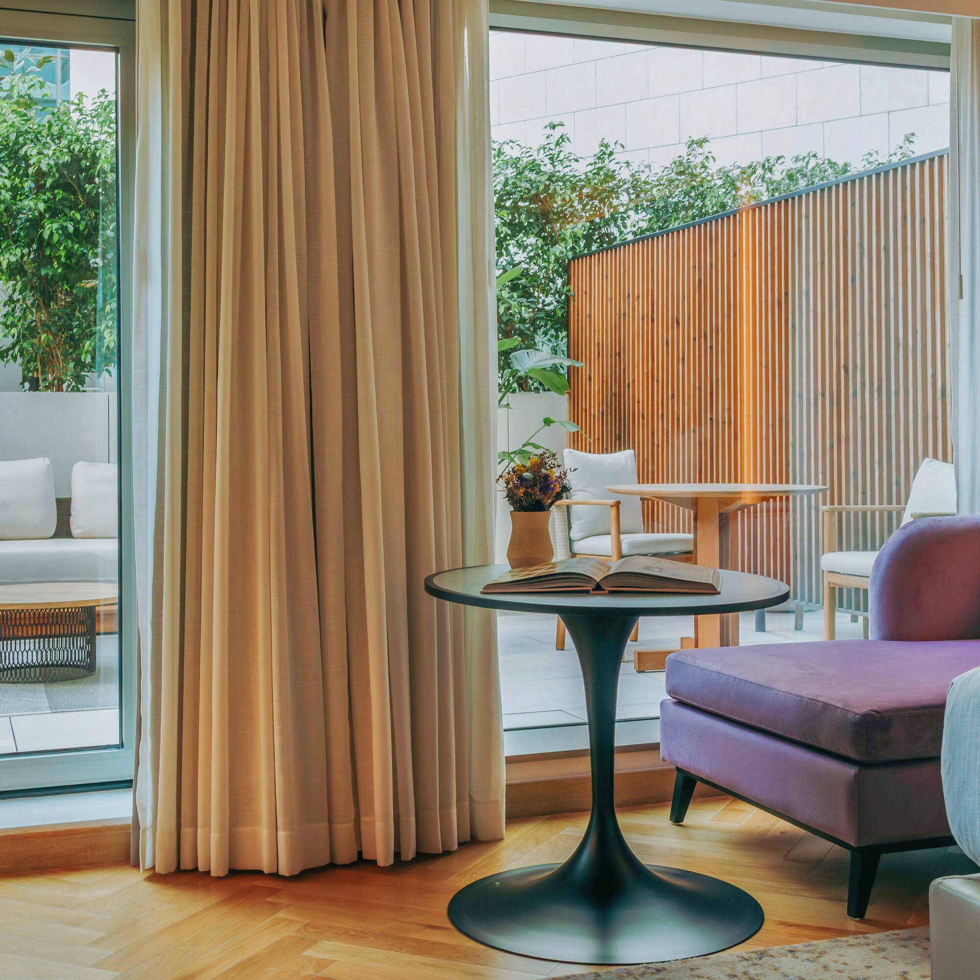 Modern hotel suite with purple sofa, outdoor patio, and lush green plants.