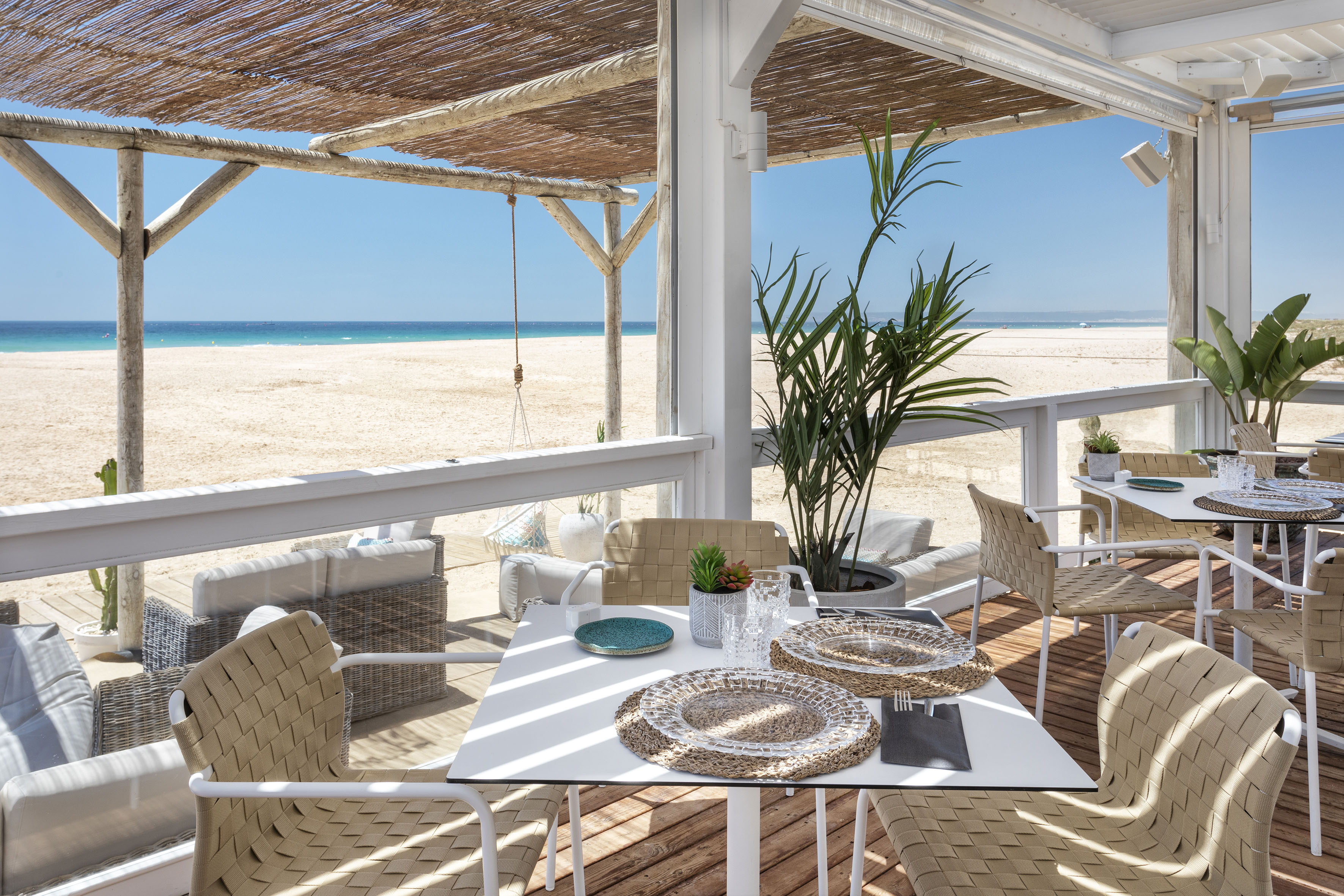 Outdoor beach restaurant with dining tables, woven chairs, and ocean view.