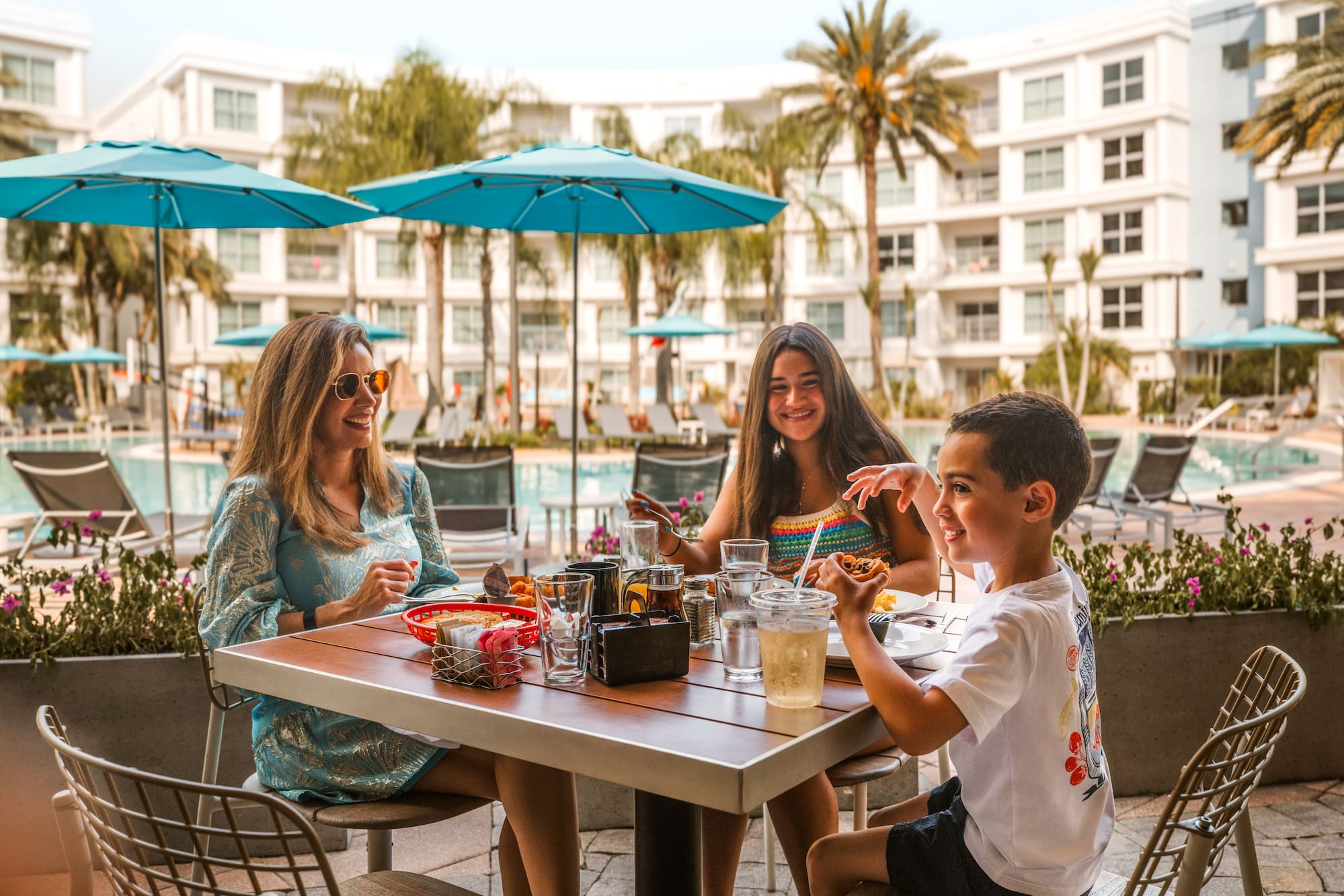 Smiling family enjoying an outdoor meal by the poolside at a tropical resort.