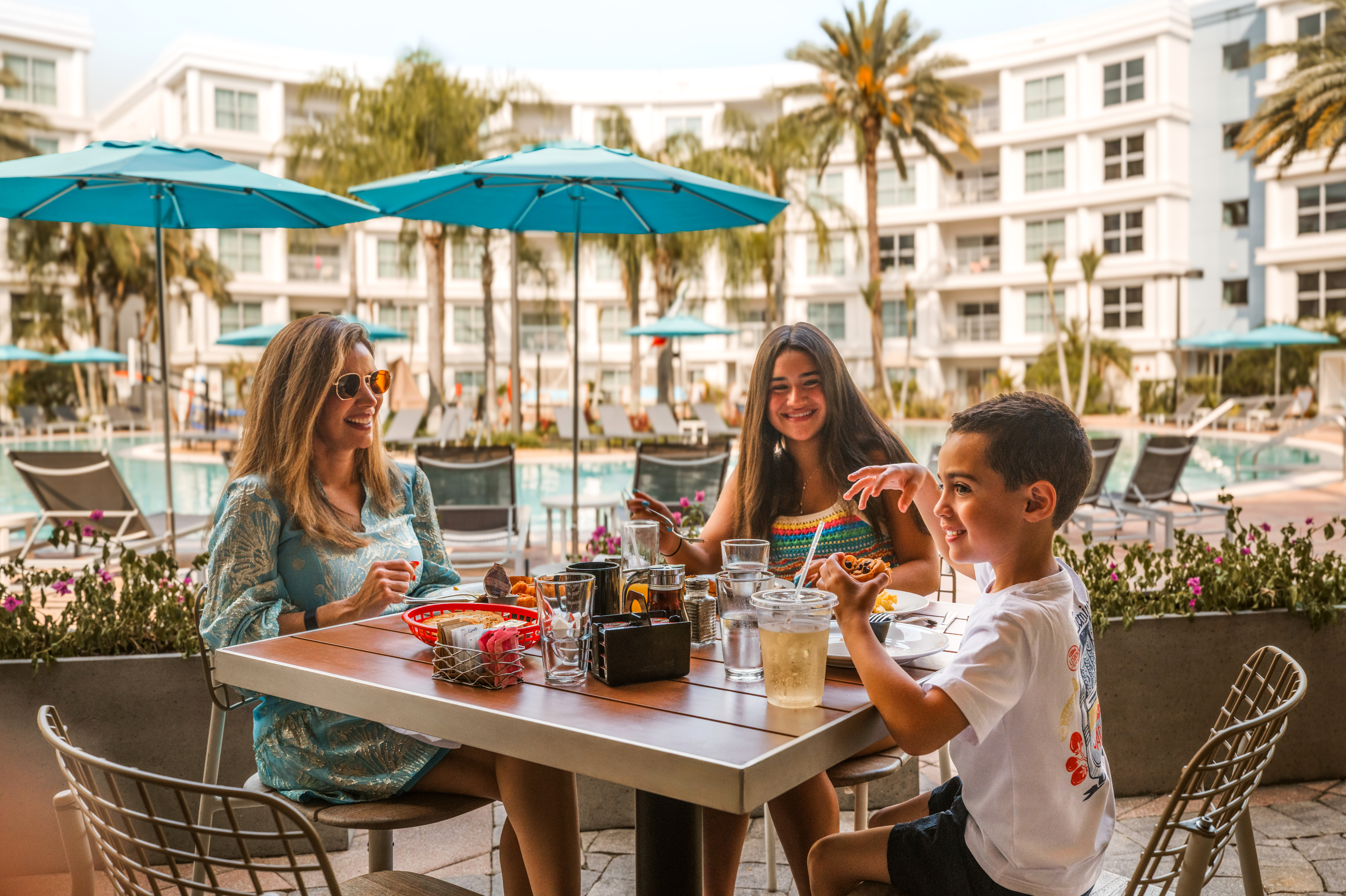 Smiling family enjoying an outdoor meal by the poolside at a tropical resort.