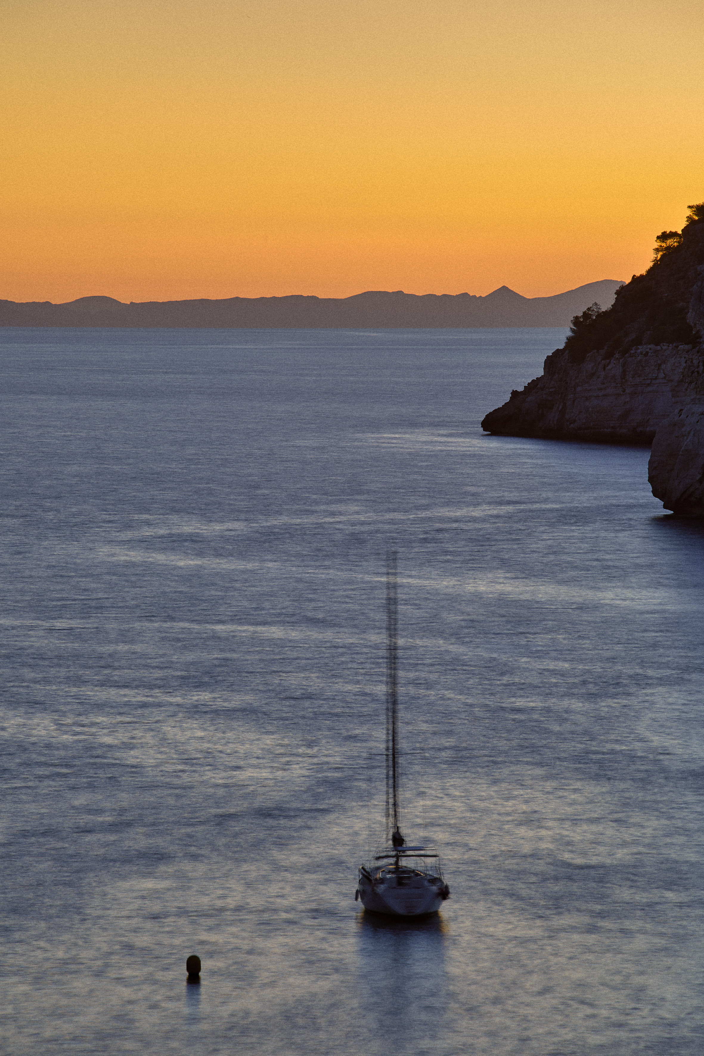 Sailboat on calm sea at sunset with orange sky and rocky coast.