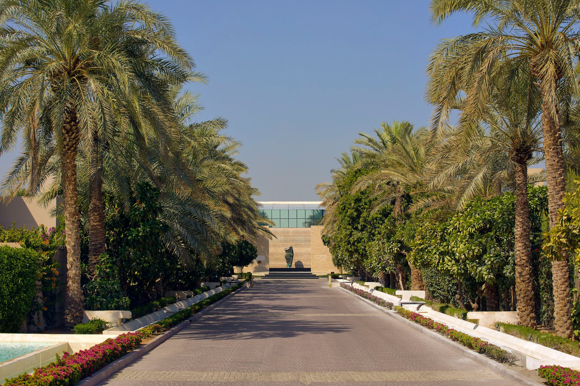 Lush palm-lined driveway leading to a modern building with central sculpture.