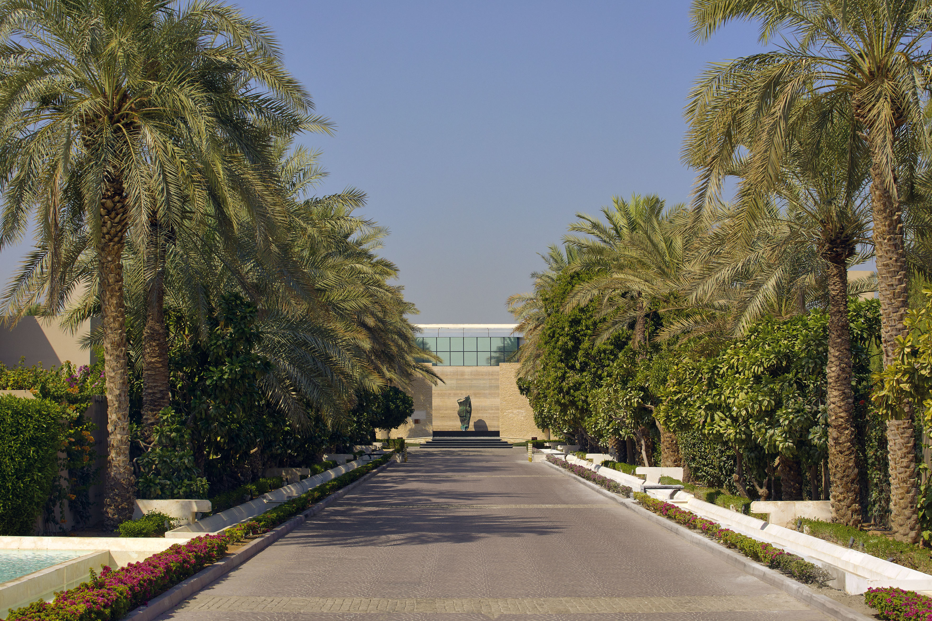 Lush palm-lined driveway leading to a modern building with central sculpture.