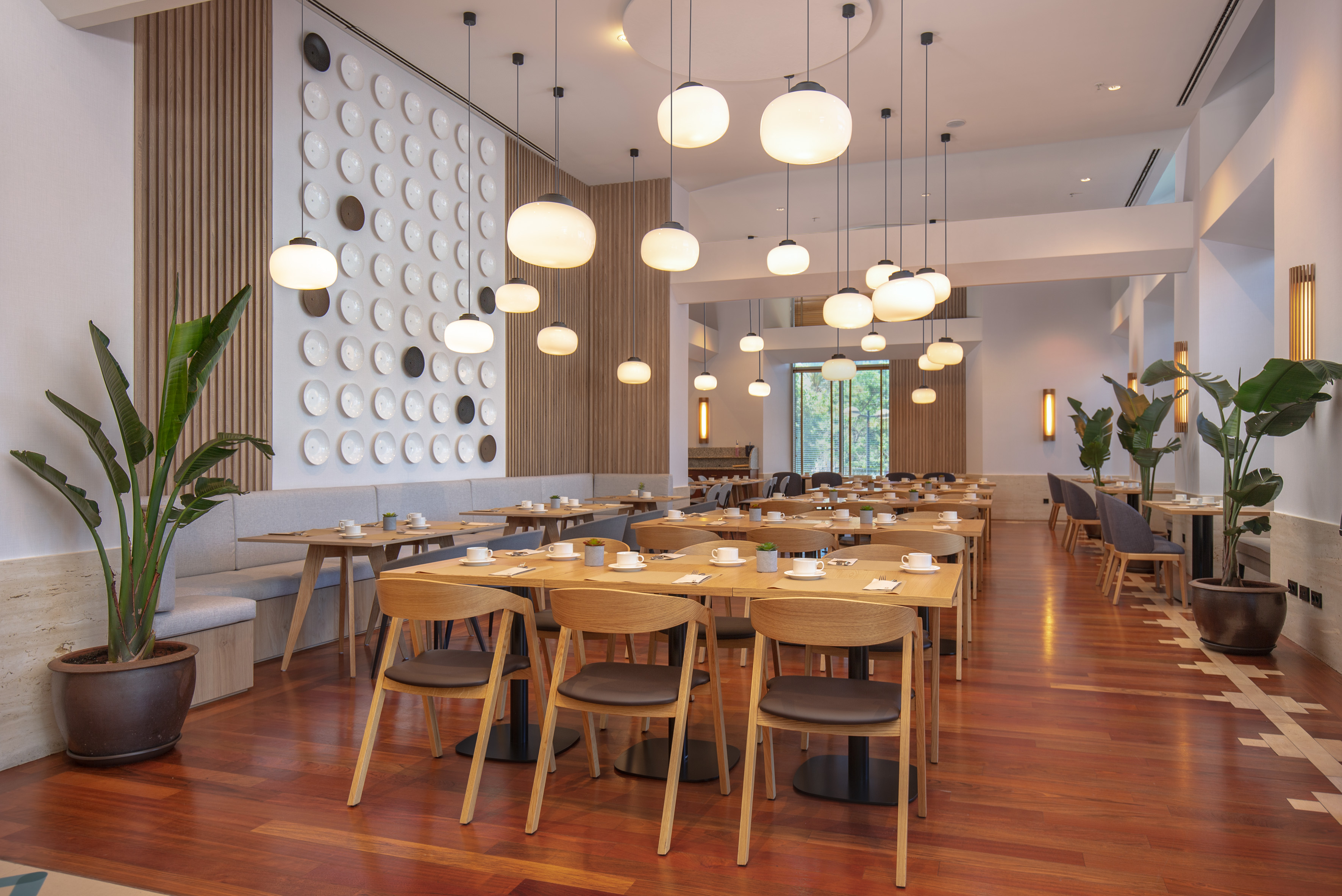 Modern restaurant dining room with wooden tables, pendant lights, and potted plants.