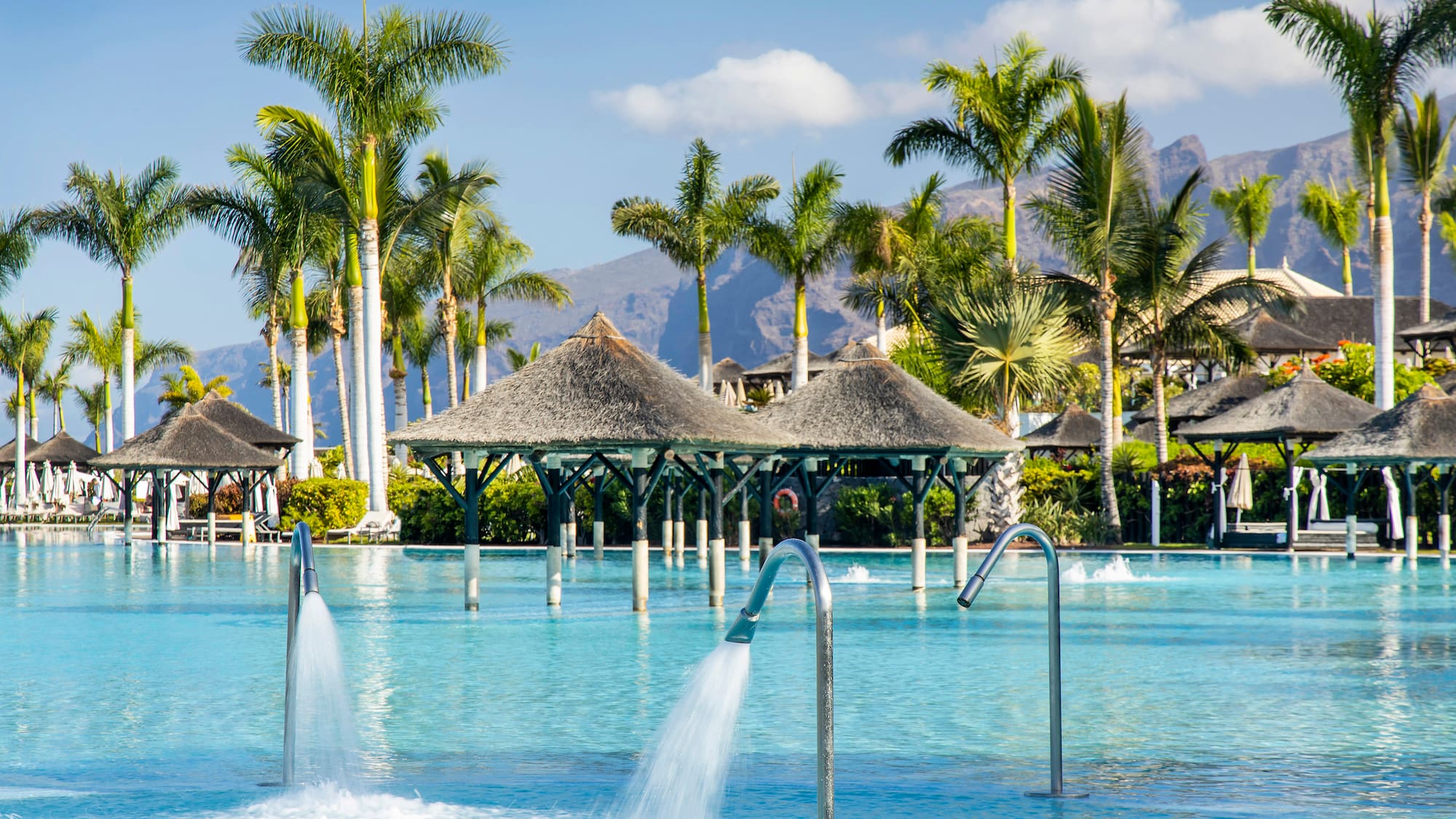 Tropical resort pool with water jets, palm trees, and distant mountains.