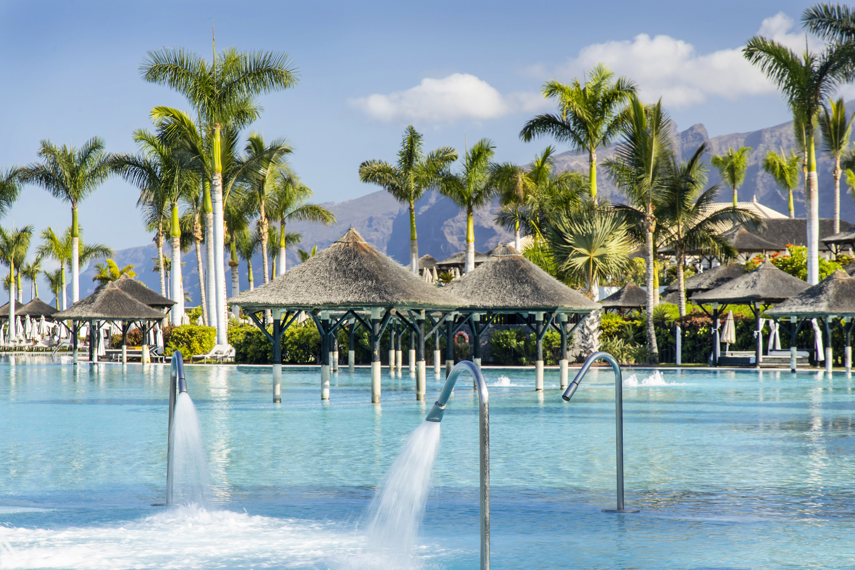 Tropical resort pool with water jets, palm trees, and distant mountains.