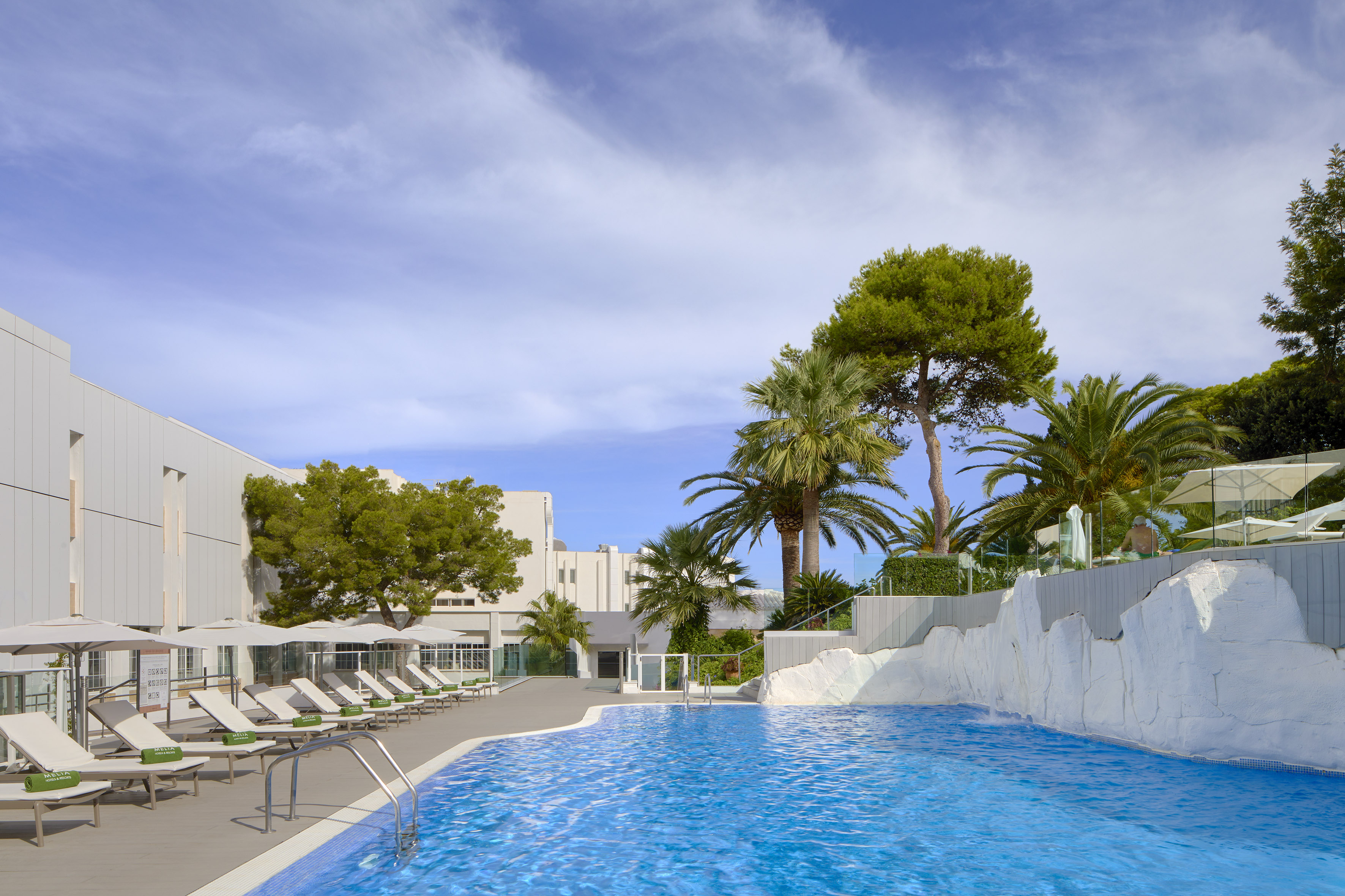 Modern resort pool with sun loungers, palm trees, and white rock feature.