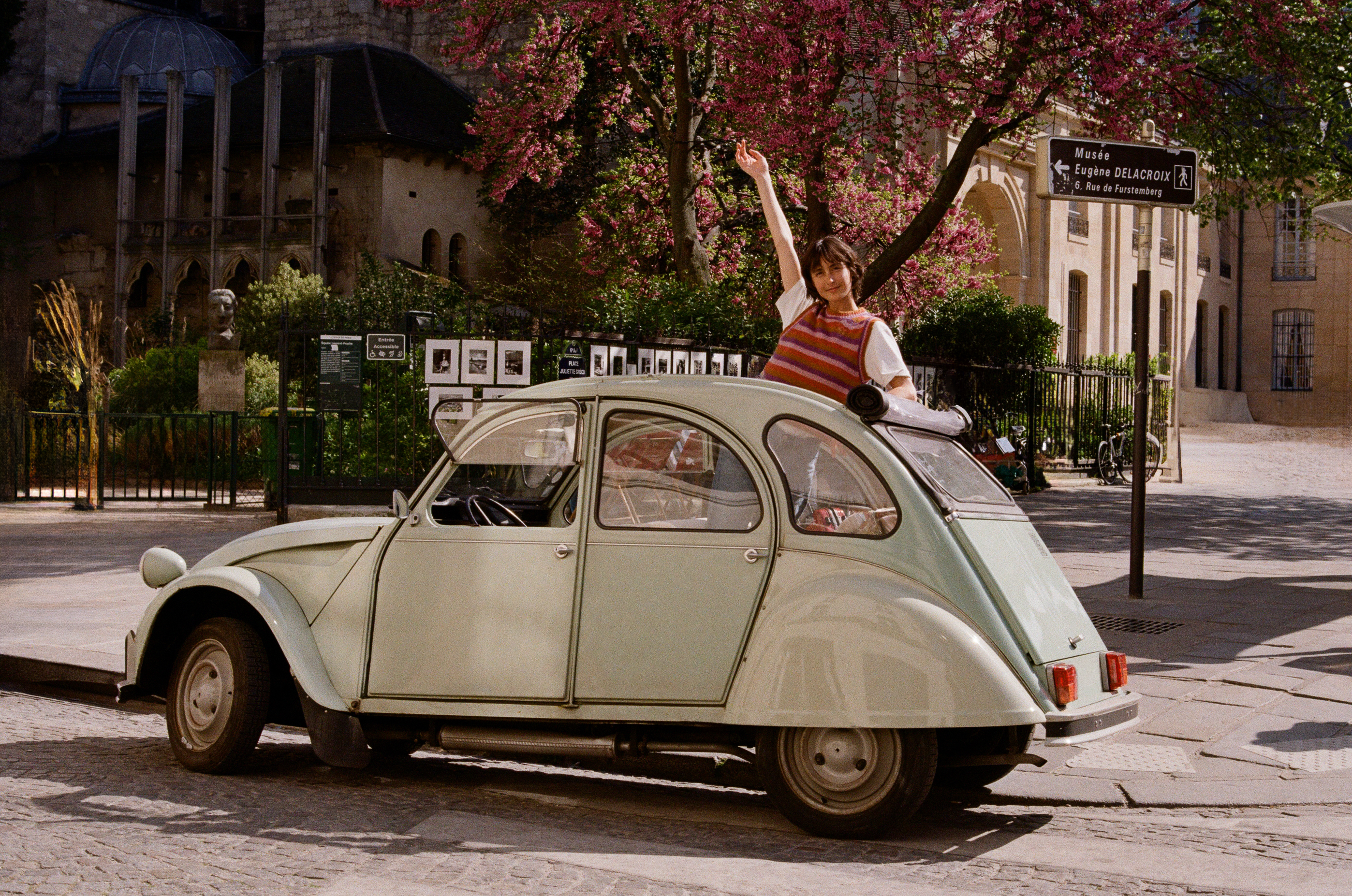 Woman waving from vintage Citroën 2CV in Paris street, blooming trees.
