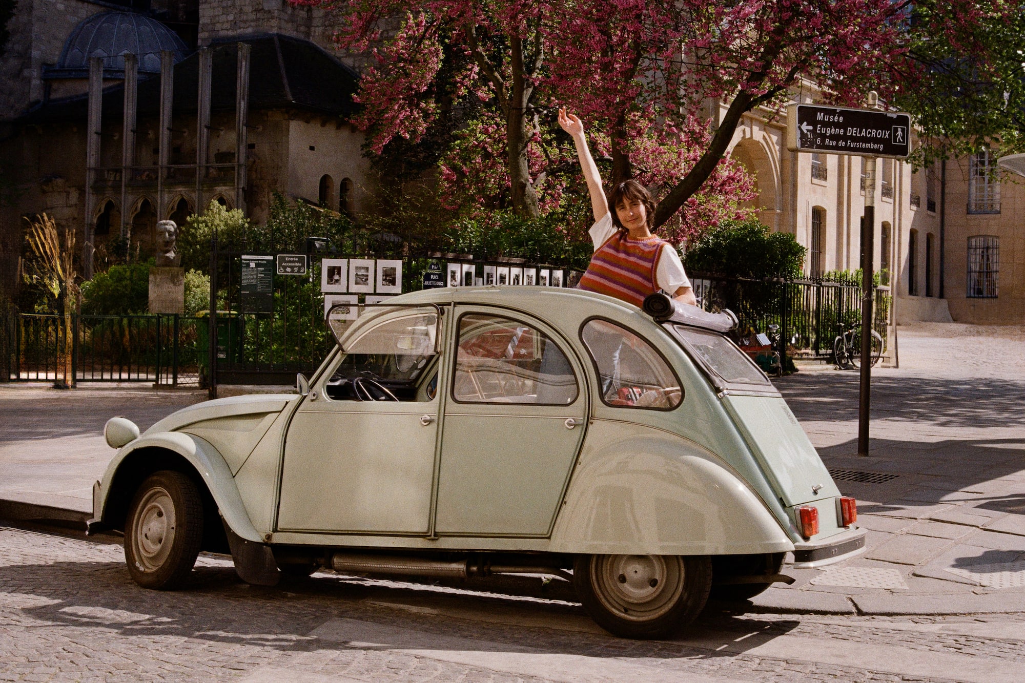 Woman waving from vintage Citroën 2CV in Paris street, blooming trees.