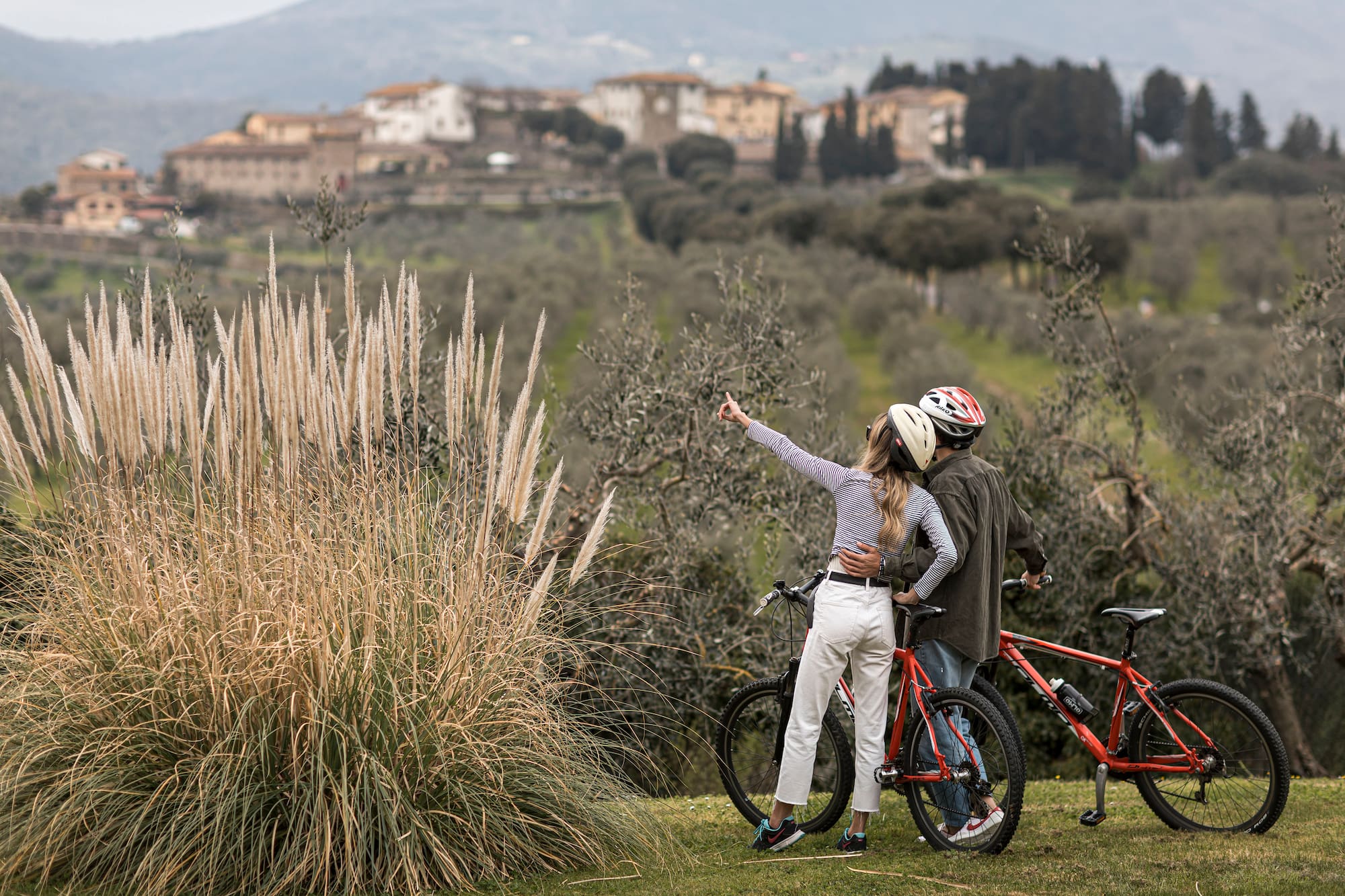 Couple on mountain bikes point to a historic Tuscan village over olive groves.