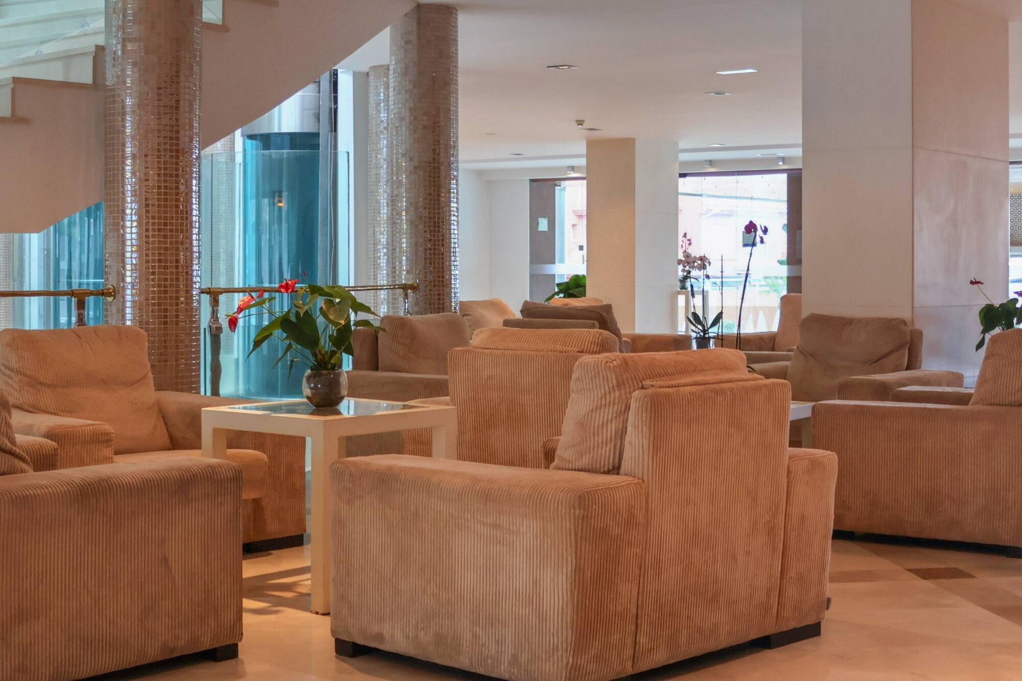 Spacious hotel lounge with beige armchairs, side tables, and a modern staircase.