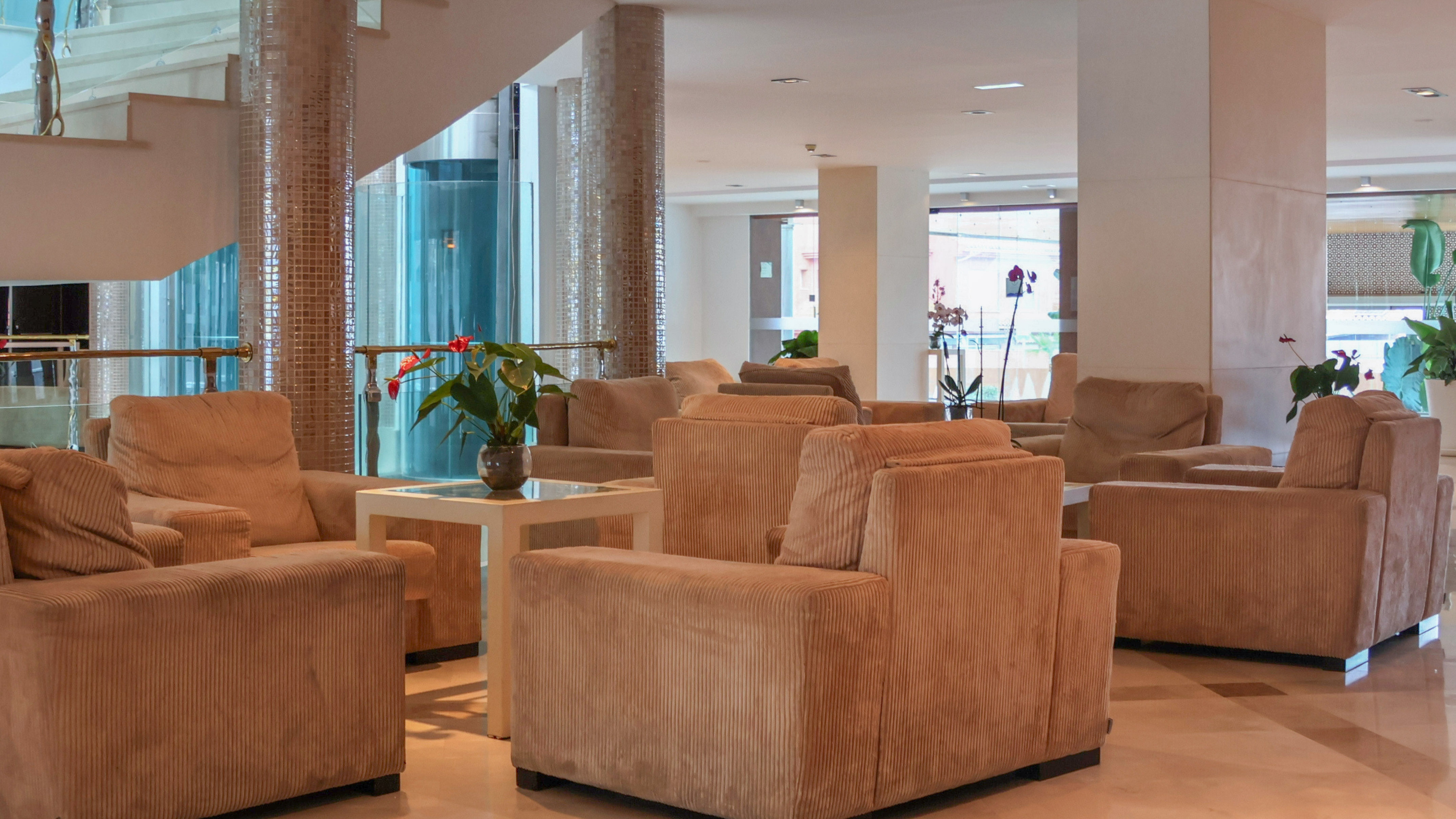 Spacious hotel lounge with beige armchairs, side tables, and a modern staircase.