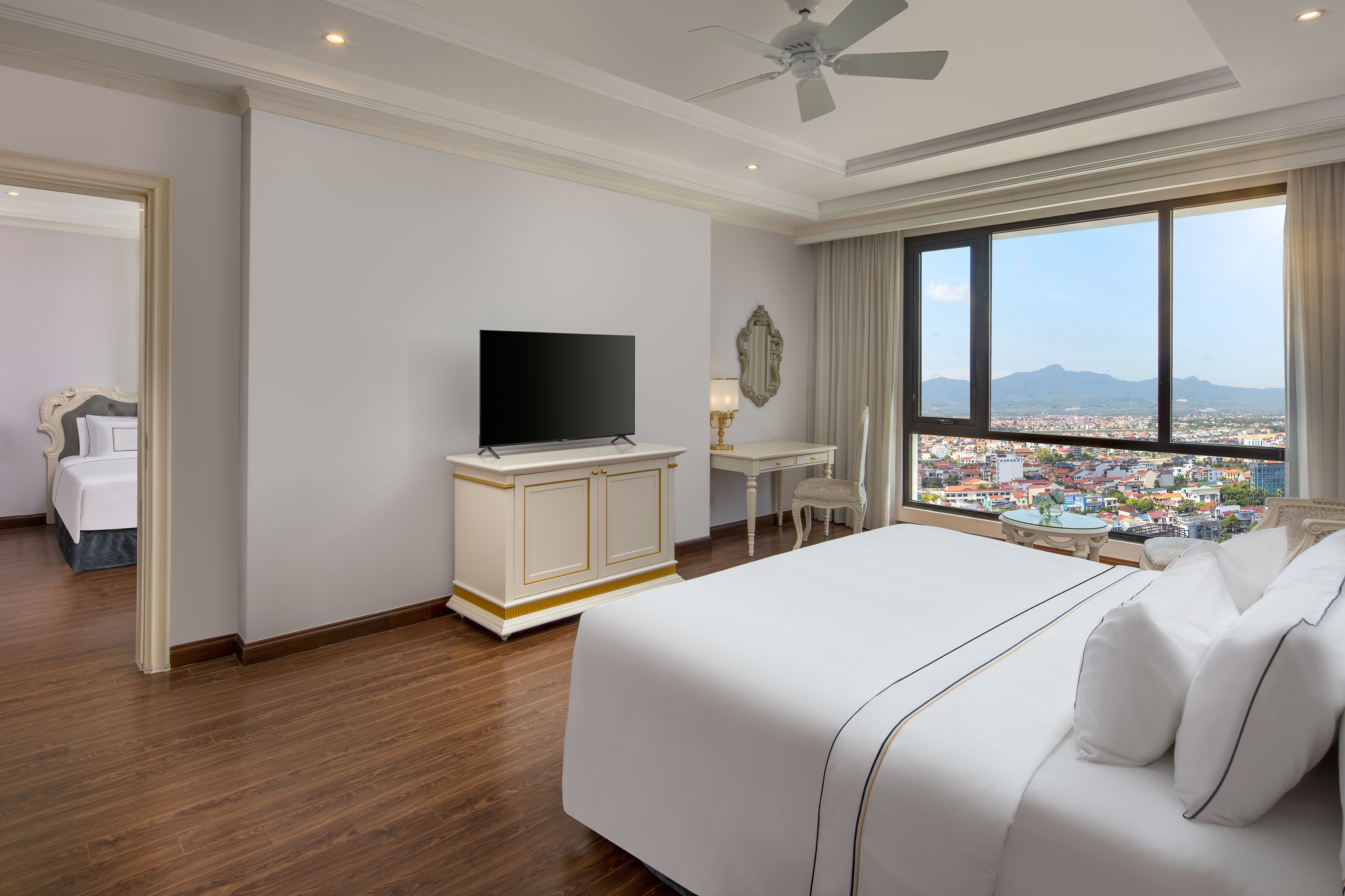 Hotel suite bedroom with city view, mountains, bed, and TV.