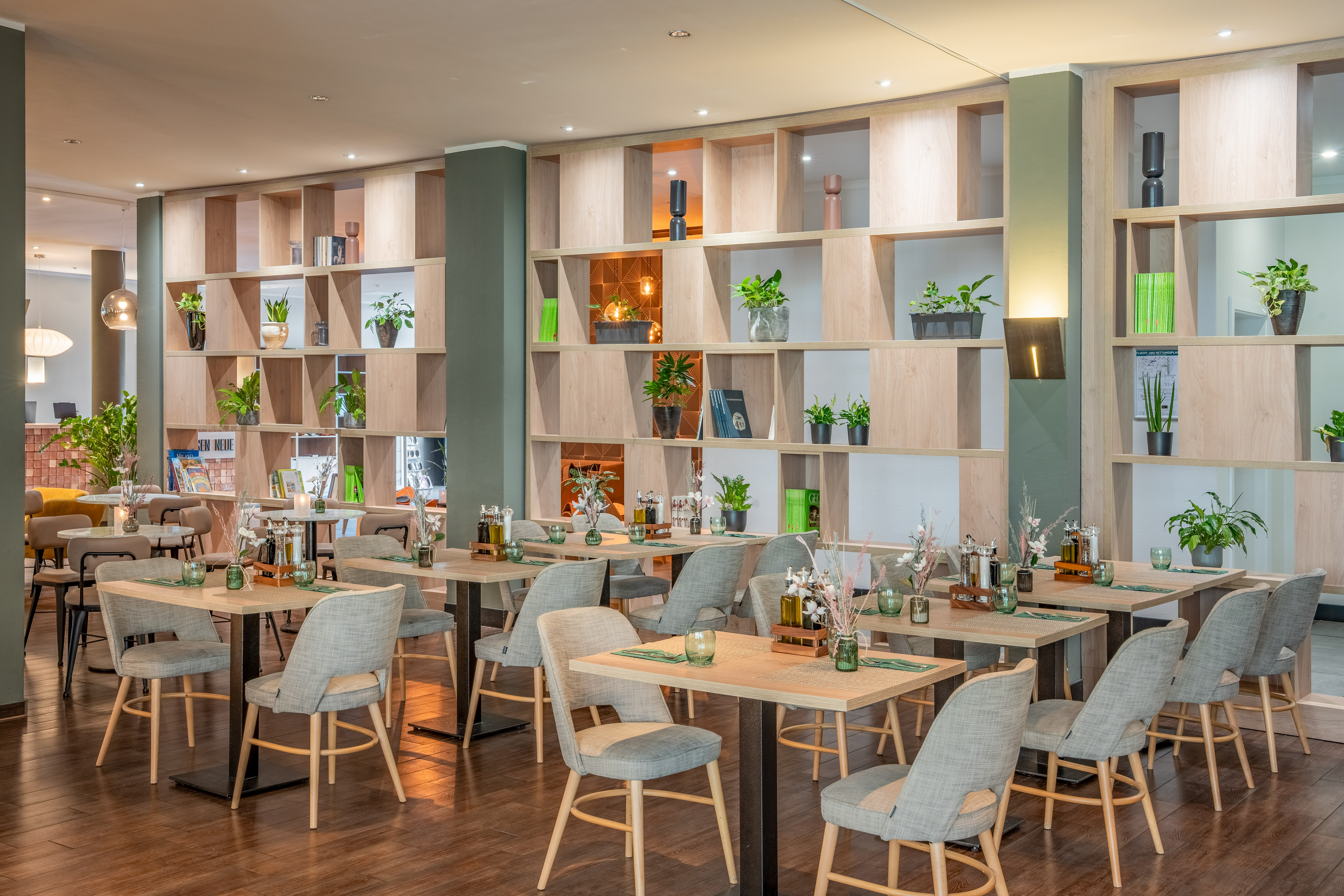 Bright restaurant interior featuring wooden tables, grey chairs, and plant shelves.