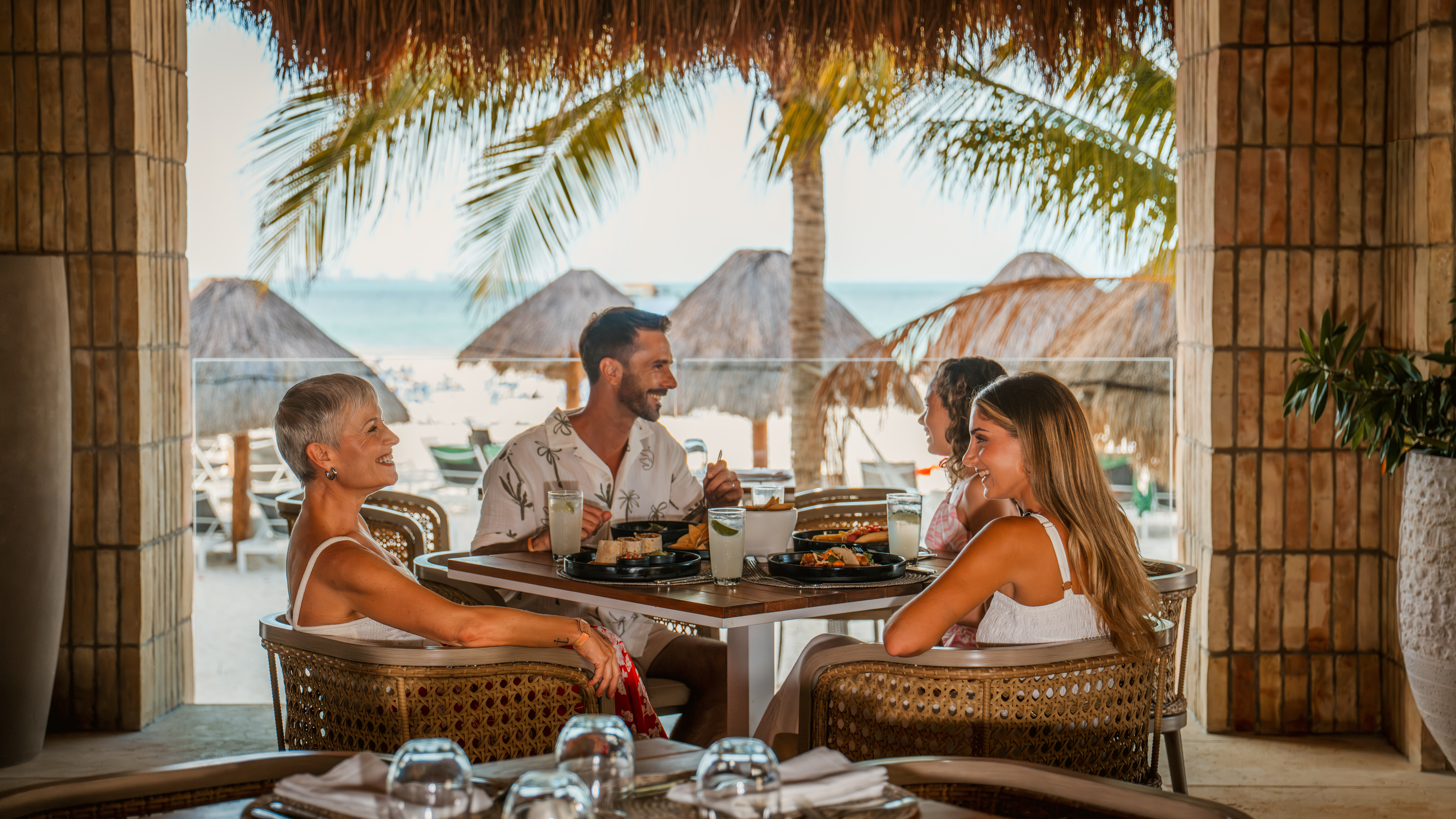 Group dining at tropical beach restaurant overlooking ocean, palapa roofs.