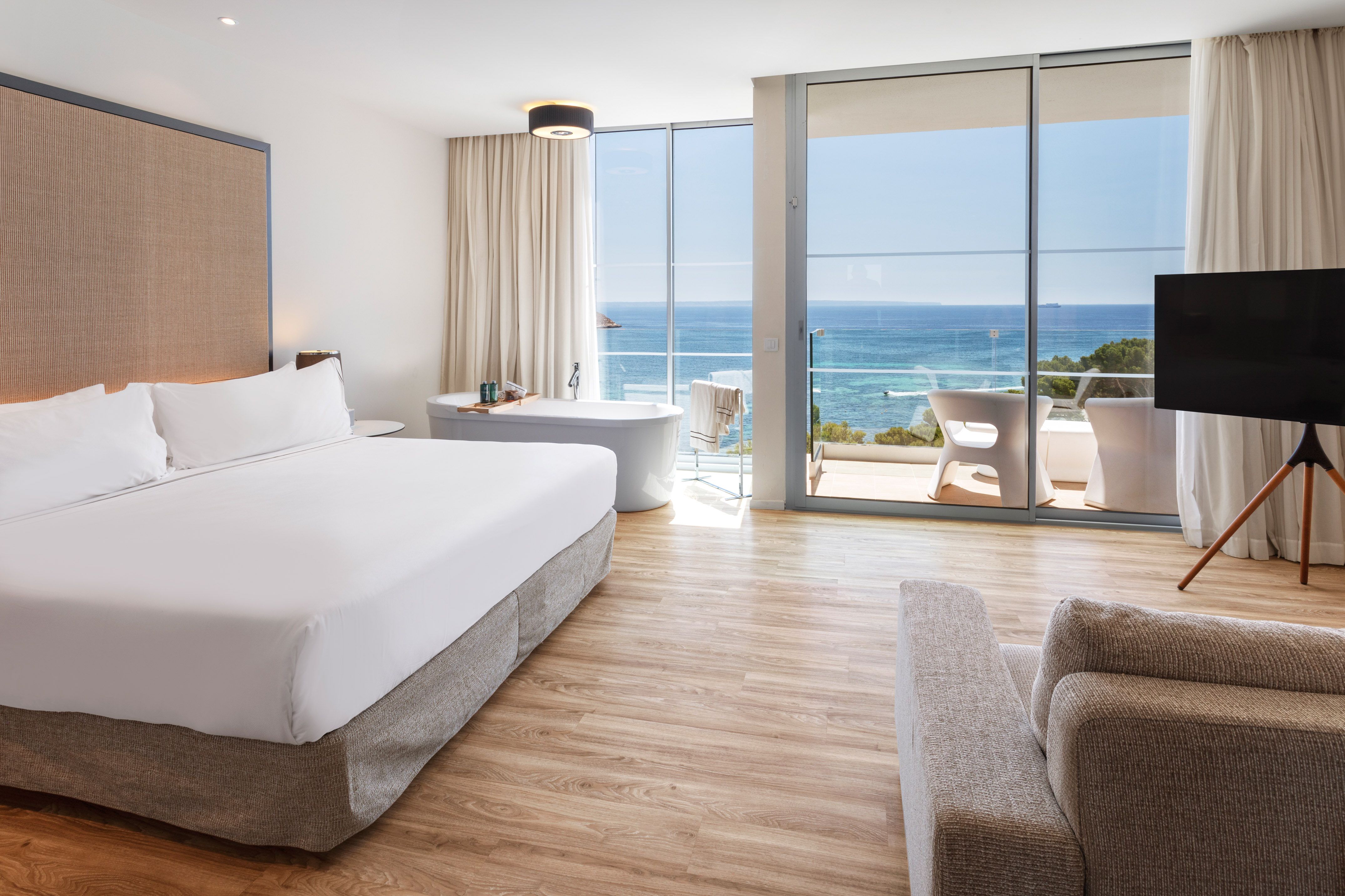 Modern luxury hotel suite with ocean view, bed, bathtub, and balcony.