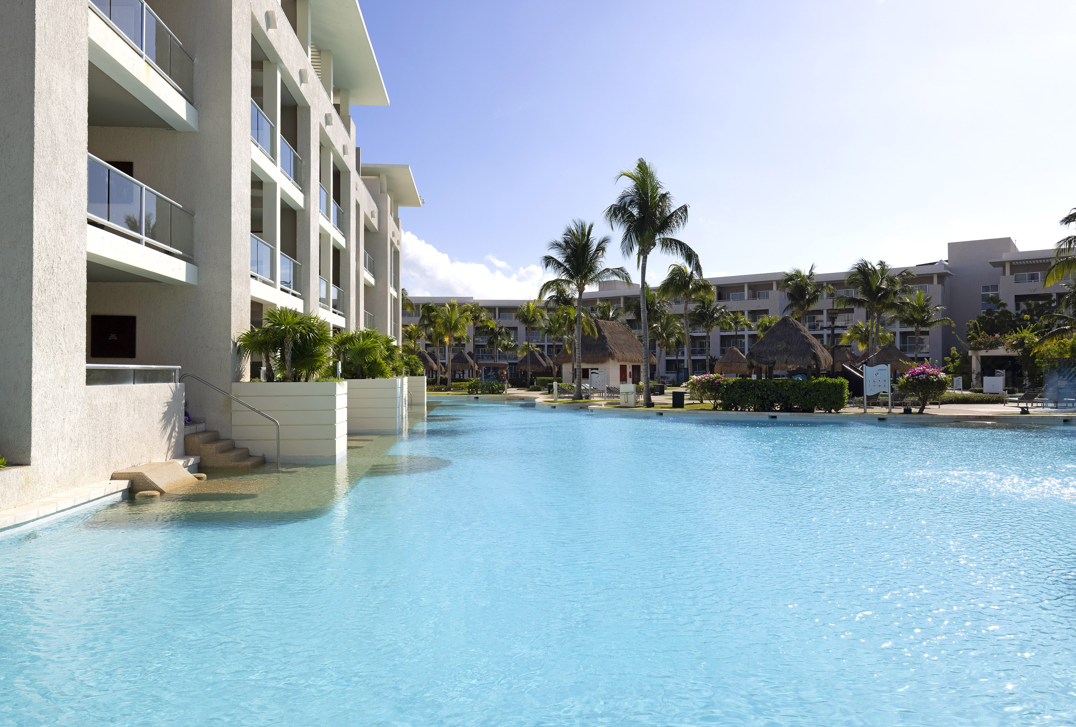 Bright blue resort pool, modern hotel, palm trees, sunny sky.