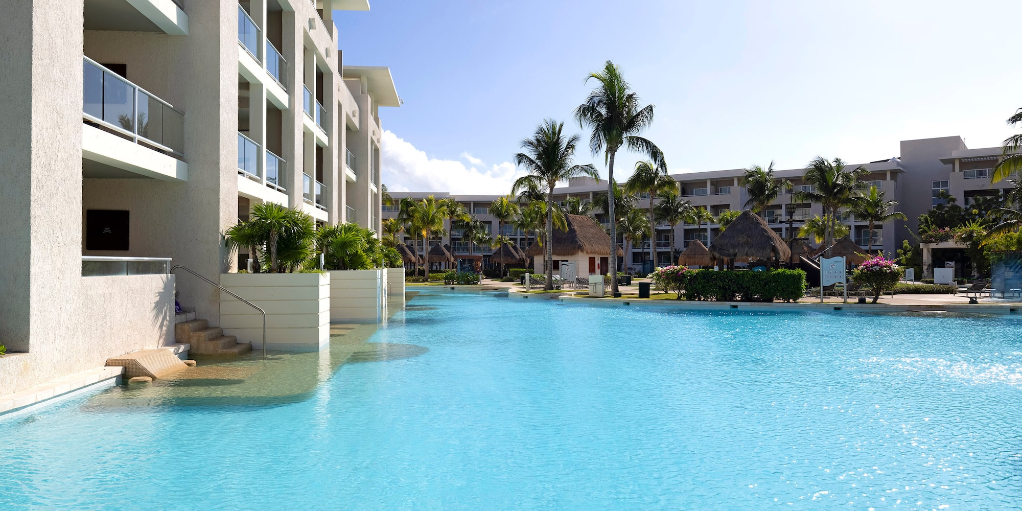 Bright blue resort pool, modern hotel, palm trees, sunny sky.