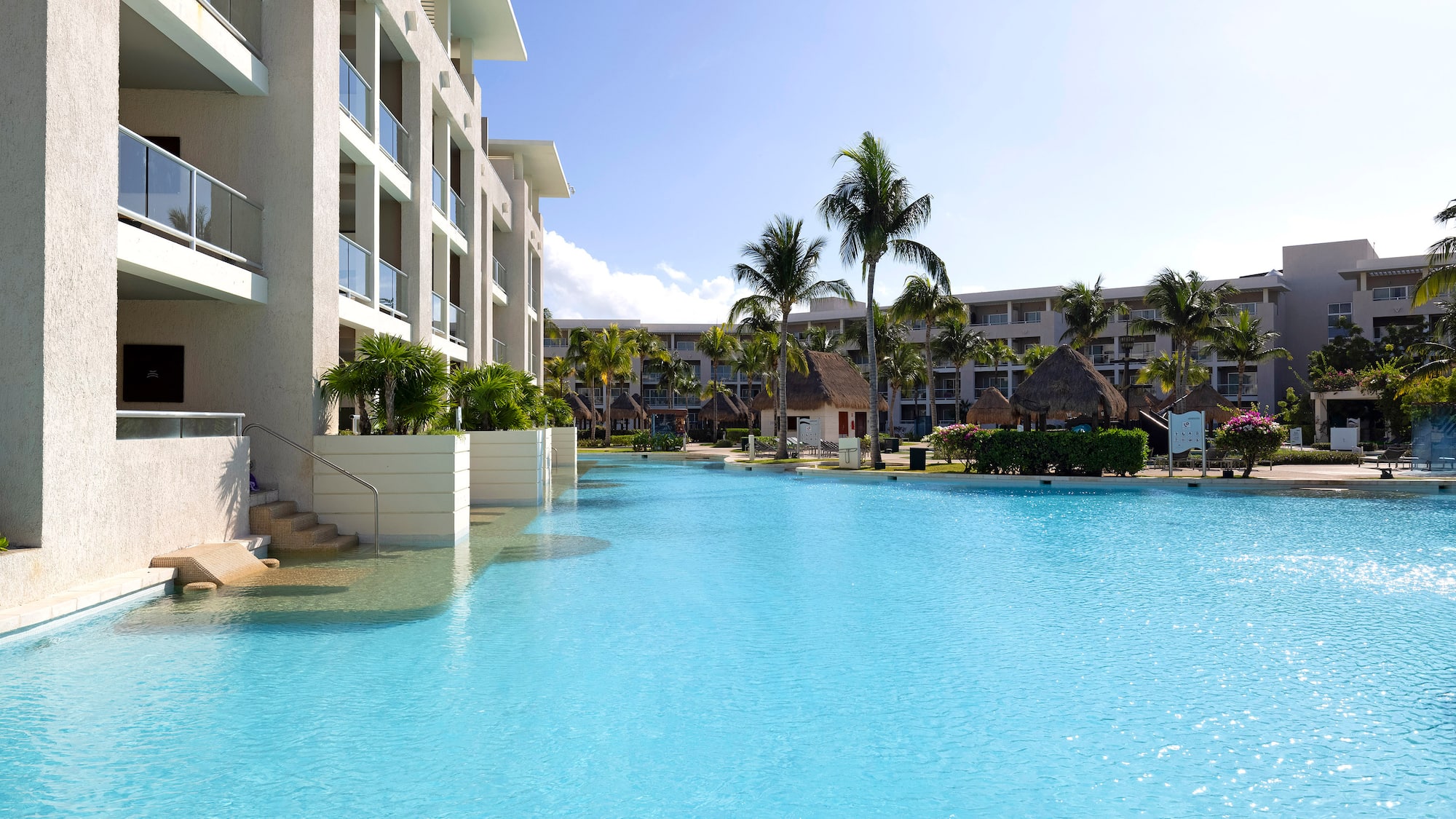 Bright blue resort pool, modern hotel, palm trees, sunny sky.