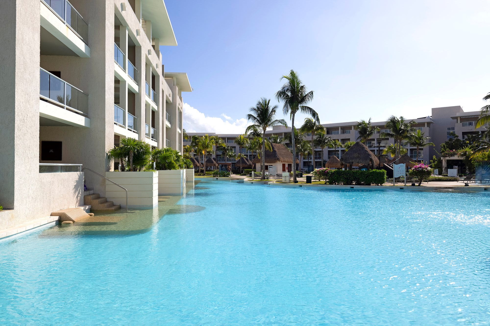 Bright blue resort pool, modern hotel, palm trees, sunny sky.