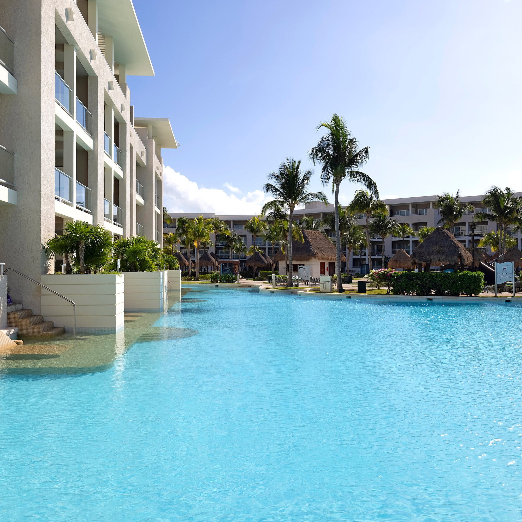 Bright blue resort pool, modern hotel, palm trees, sunny sky.