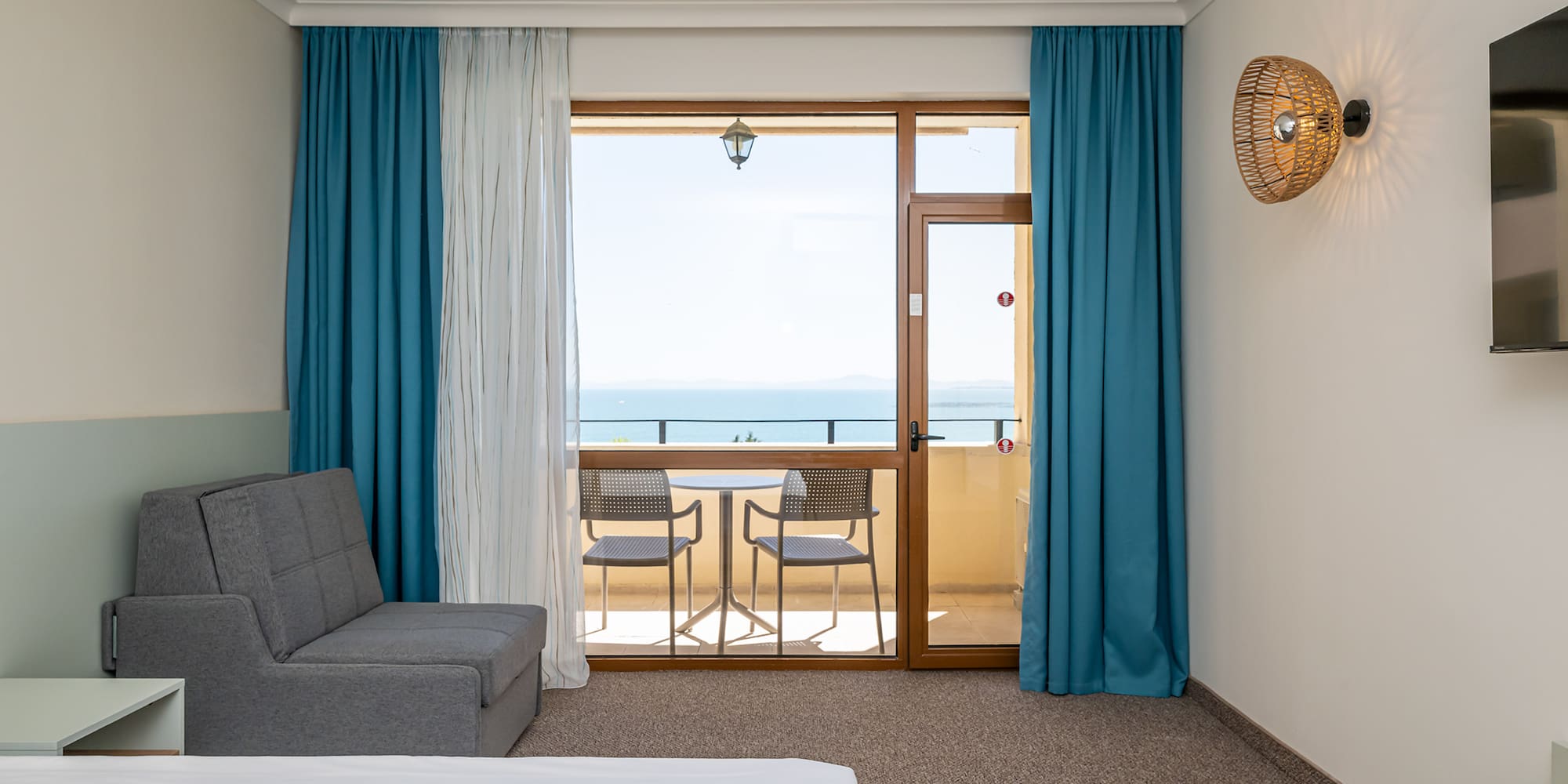 Hotel room with sea view balcony, gray sofa bed, and blue curtains.