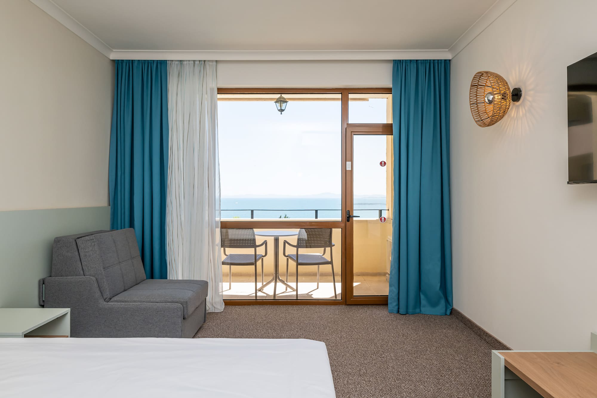 Hotel room with sea view balcony, gray sofa bed, and blue curtains.