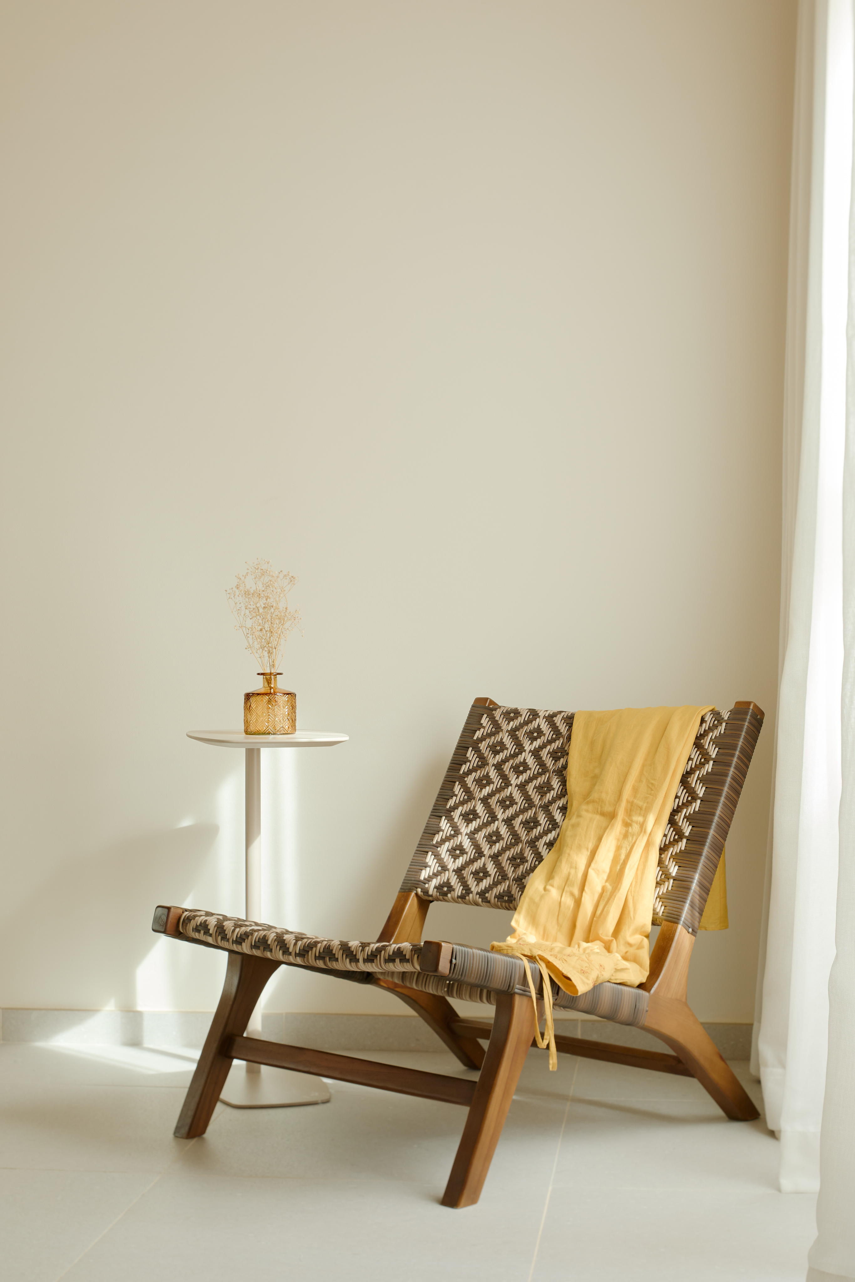 Woven lounge chair, yellow throw, dried flowers on side table in sunny room.