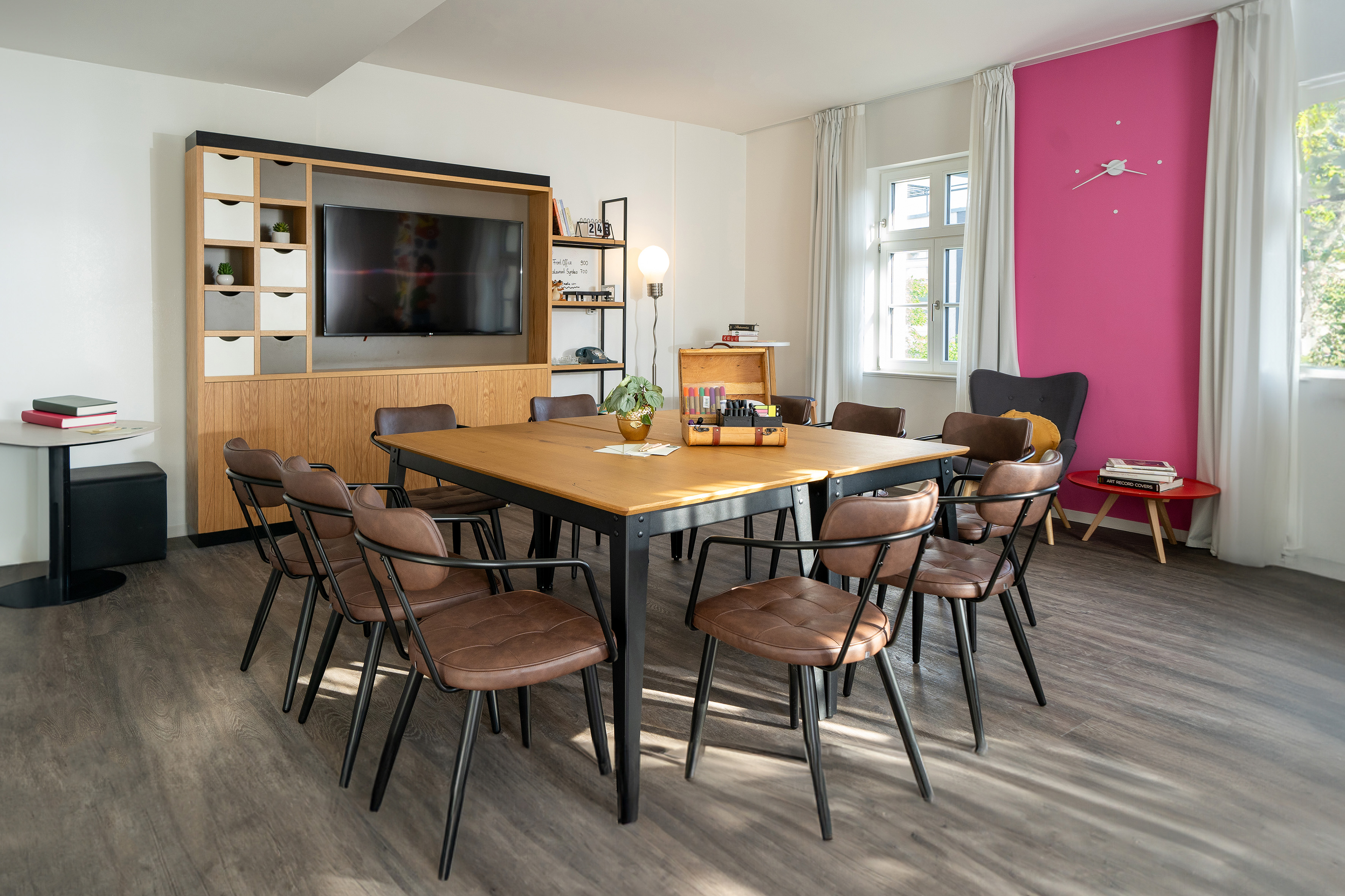 Modern meeting room with wooden table, brown chairs, TV, and a bright pink accent wall.